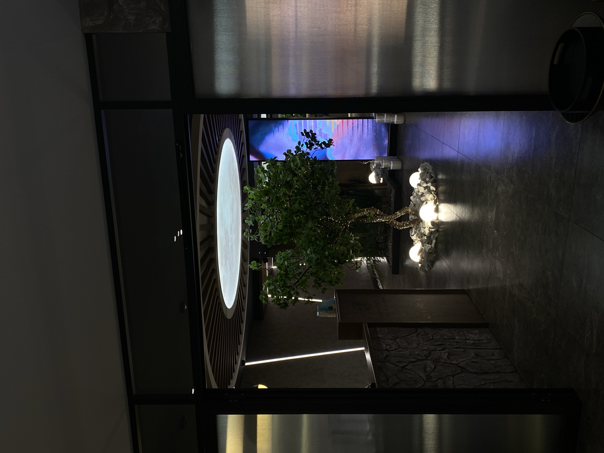 The image captures an indoor hallway or entryway, looking into a modern, dimly lit interior space. On the left, a large, prominent circular light feature is embedded in a dark, slatted wooden wall, casting a soft, ambient glow. Centrally positioned within the frame is a decorative artificial tree, adorned with small, twinkling lights, providing a touch of nature and sparkle.
Behind the tree, a large rectangular screen displays vibrant abstract art, predominantly featuring hues of blue, purple, and pink, which adds a dynamic and colorful backdrop. To the right of the tree, a cluster of spherical white lights are arranged on the floor amidst white, rocky decorative elements. The floor throughout the visible area is dark grey tile, exhibiting slight reflections of the various light sources.
The walls are dark, with some sections featuring a textured, stone-like finish. A thin, linear LED light strip runs diagonally across the lower left wall or floor, adding to the contemporary lighting design. The overall scene is illuminated entirely by artificial lights, suggesting an evening setting or a space designed to maintain a consistent ambiance regardless of natural light. The entrance to this area is framed by dark, sleek structures, possibly metal. In the upper right corner, a dark, circular object, perhaps a decorative planter or tray, is visible. No people are present, and no discernible text is visible within the image. The atmosphere is modern, serene, and sophisticated.