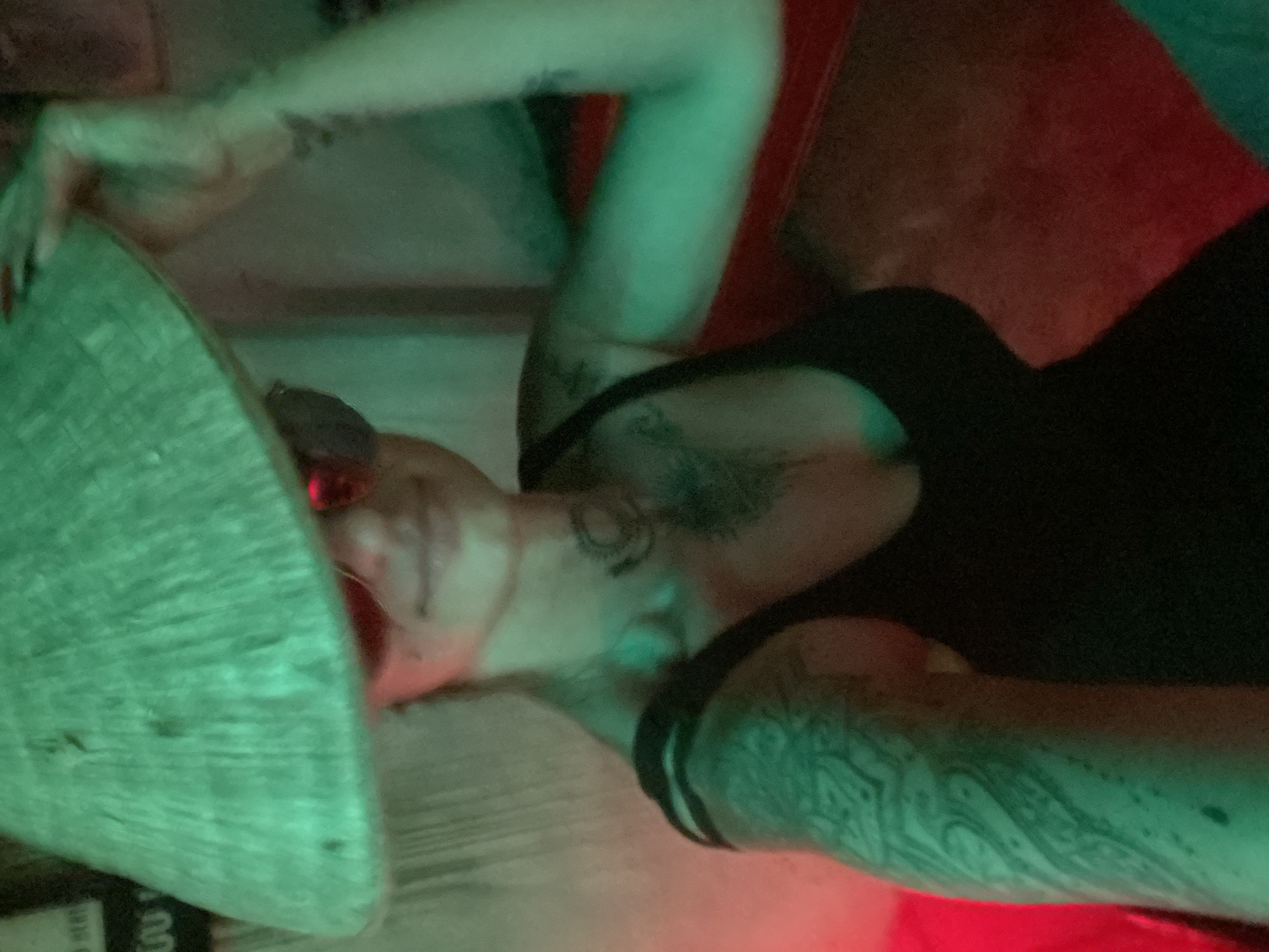A person lies on their back, wearing sunglasses and a conical Asian hat. They are mostly dressed in black and have tattoos on their arms and chest. The scene is illuminated by green and red lighting. The location is potentially Mũi Né, Vietnam.