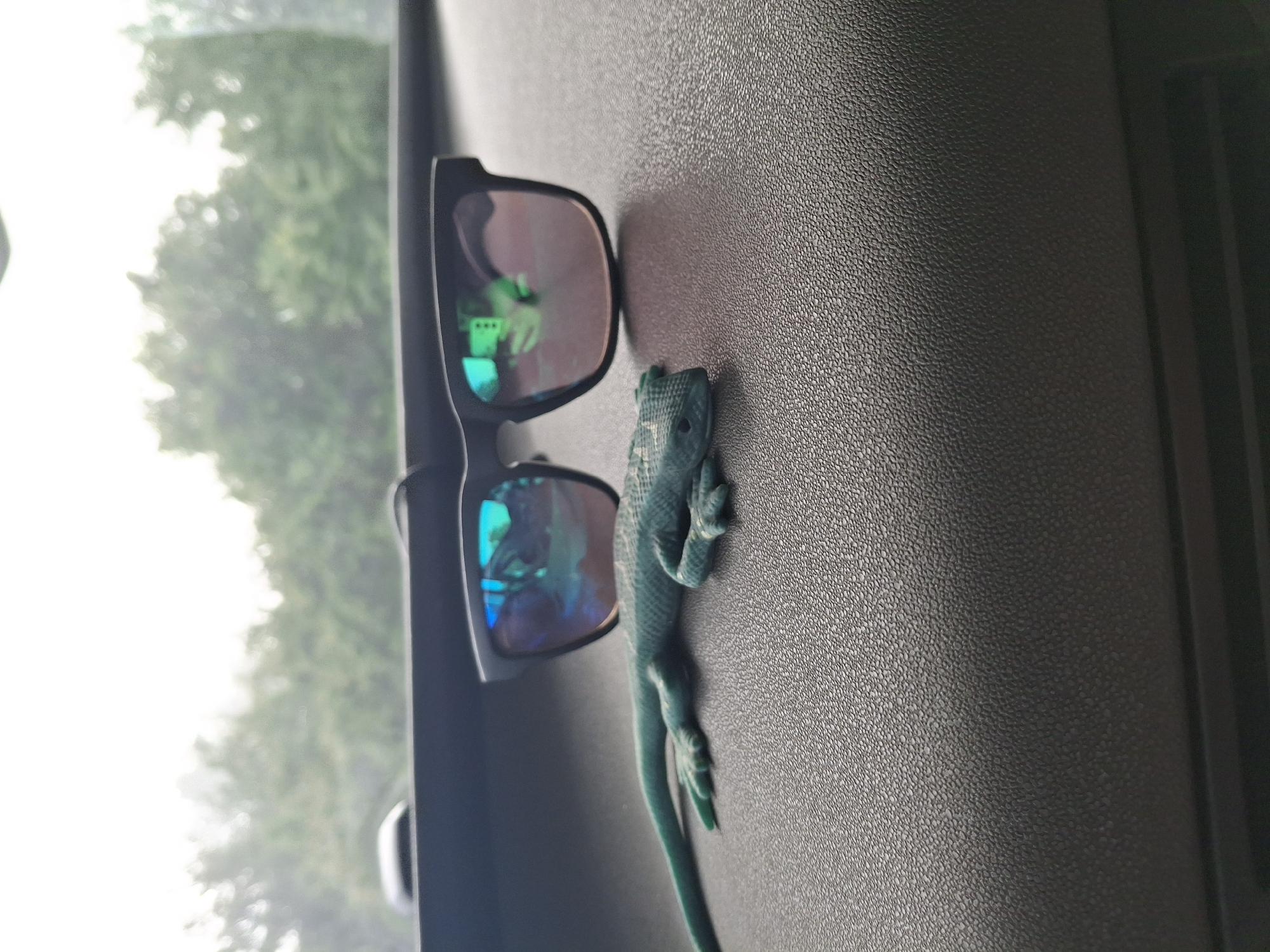 An indoor shot captured from within a vehicle shows a pair of dark sunglasses and a small green lizard (possibly a toy or model) resting on a textured, dark gray surface, likely a car dashboard or side panel. The image is oriented vertically, with the subjects appearing on the right side of the frame.
The sunglasses, with black frames, have reflective, reddish-orange tinted lenses that show bright green and blue reflections, likely from the outdoor environment visible through the vehicle's window. The lizard, a vibrant green, is positioned to the right of the sunglasses, facing towards the top of the frame. Its skin has a visible texture.
To the left of the subjects, a bright, diffused light source dominates the background, indicating an outdoor scene through a window. Blurry green foliage, consistent with trees or bushes, is visible against the bright sky, suggesting daytime. The weather appears to be bright, possibly overcast, given the lack of direct shadows and the diffuse light. The dark gray surface on which the items rest has a finely pebbled texture. No discernible activity is taking place; the items are static. There is no visible text in the image. The location context is Osikovitsa, Bulgaria.