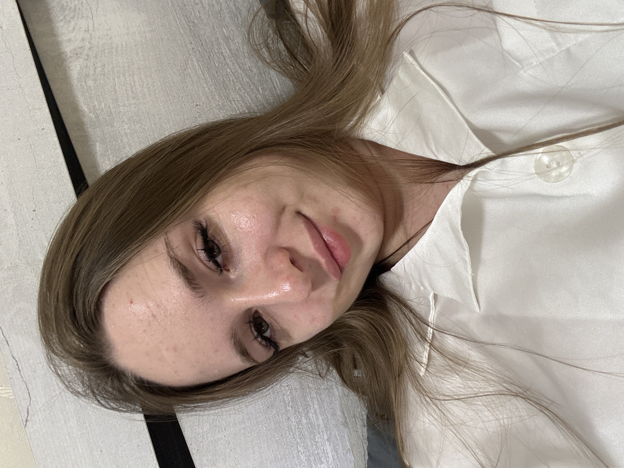 A young Caucasian woman with light brown hair is captured in a close-up selfie, lying down. Her head is tilted slightly, and she is looking directly at the camera with a subtle smile. Her makeup includes noticeable eyeliner and mascara, and her lips are painted with a glossy pink lipstick. The background appears to be a wall with a textured pattern and a dark horizontal line. She is wearing a white collared shirt. The image quality is slightly grainy, suggesting it might have been taken in low light conditions.