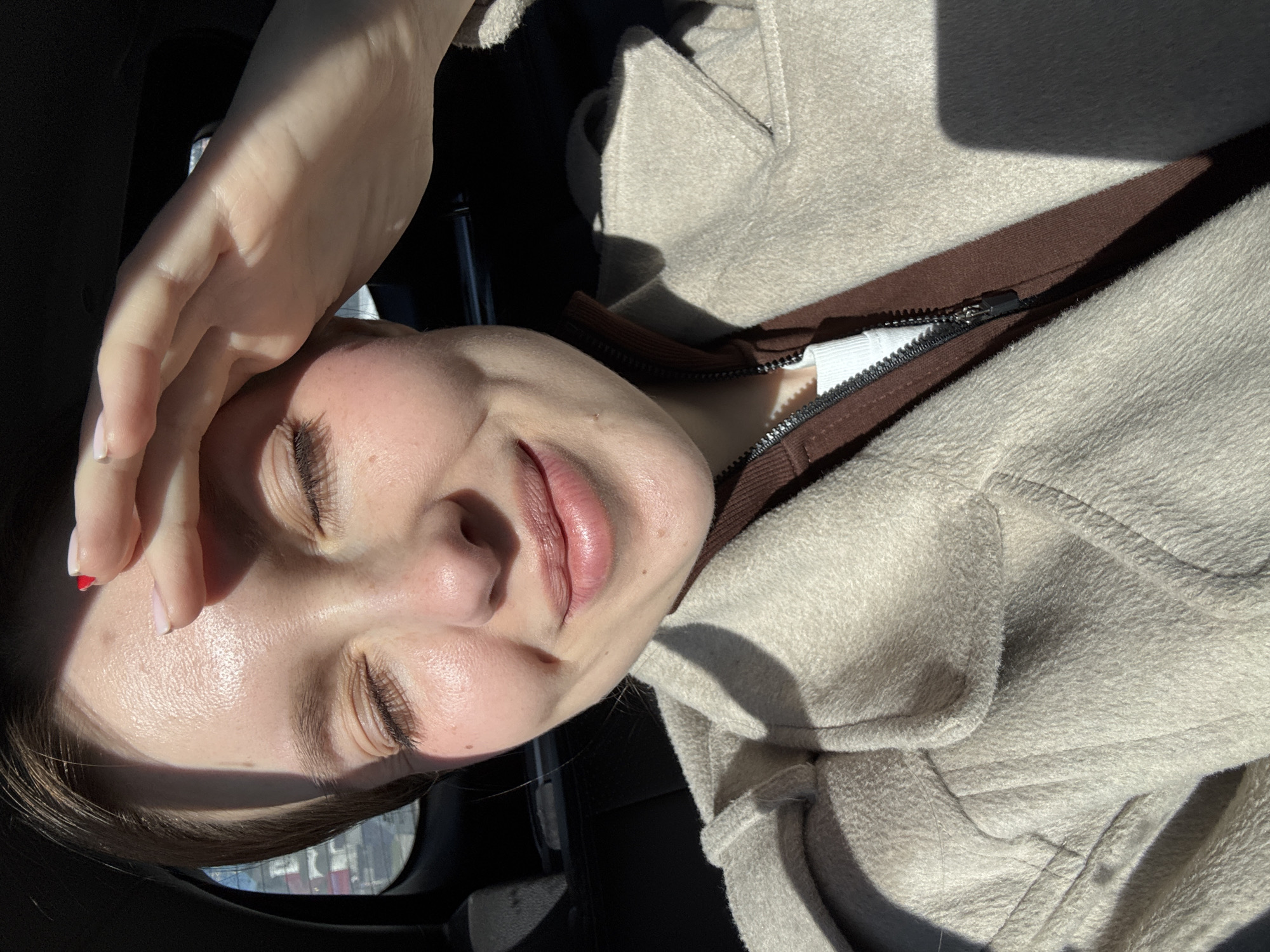 A woman is shielding her eyes from the sun with her hand. She has fair skin, brown hair, and closed eyes. She is wearing a beige jacket with a brown trim and a white top underneath. The background is dark, suggesting she is inside a vehicle. The reflection in the car window shows buildings, suggesting an urban environment, potentially Moscow, Russia.