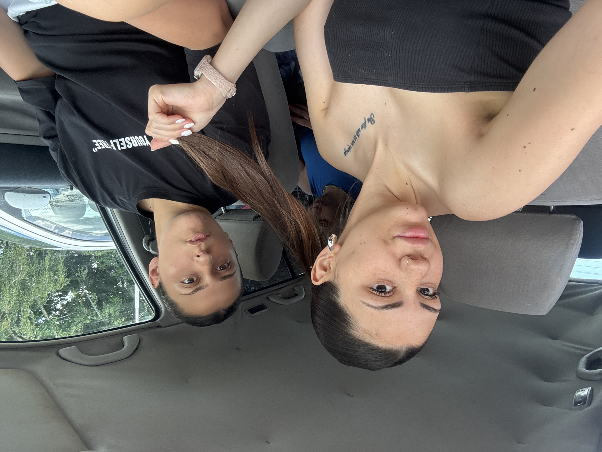 The image depicts three individuals inside a parked vehicle, identified as being in the city of Sofia, Bulgaria, during daylight hours. In the foreground, a young woman is seated in what appears to be the front passenger seat. She has dark hair pulled back, a light complexion with visible freckles on her nose and cheeks. She wears a small silver hoop nose ring on her right nostril and a large silver hoop earring in her left ear. Her attire consists of a black strapless top. A tattoo reading "She flies with her own wings" is visible on her upper left chest/collarbone area. Her right hand, featuring white nail polish, holds a strand of long, dark hair belonging to the individual next to her. She is looking directly towards the camera with a subtle expression.
To her right, a second woman is seated, likely in the driver's seat. She has dark hair pulled back into a ponytail, a slightly darker complexion, and a neutral expression as she also looks towards the camera. She wears a black oversized t-shirt with white text partially visible on the front, reading "...YOURSELF FREE". Her right hand is holding a section of her own dark brown ponytail. A light pink silicone wristband or smartwatch is present on her right wrist.
A third individual is partially visible in the back seat, positioned centrally between the two front occupants. This person has lighter-colored hair, possibly blonde or light brown, and is wearing a blue garment. Only the top of their head and a portion of their upper body are discernible.
The vehicle's interior features a gray fabric headrest for the front passenger seat, and a gray textured headliner on the roof with visible grab handles and some minor indentations. Through the windows, dense green foliage, consistent with trees, is evident, indicating an outdoor environment. A portion of another vehicle, possibly silver or light blue, and a white pole are visible through the driver's side window.