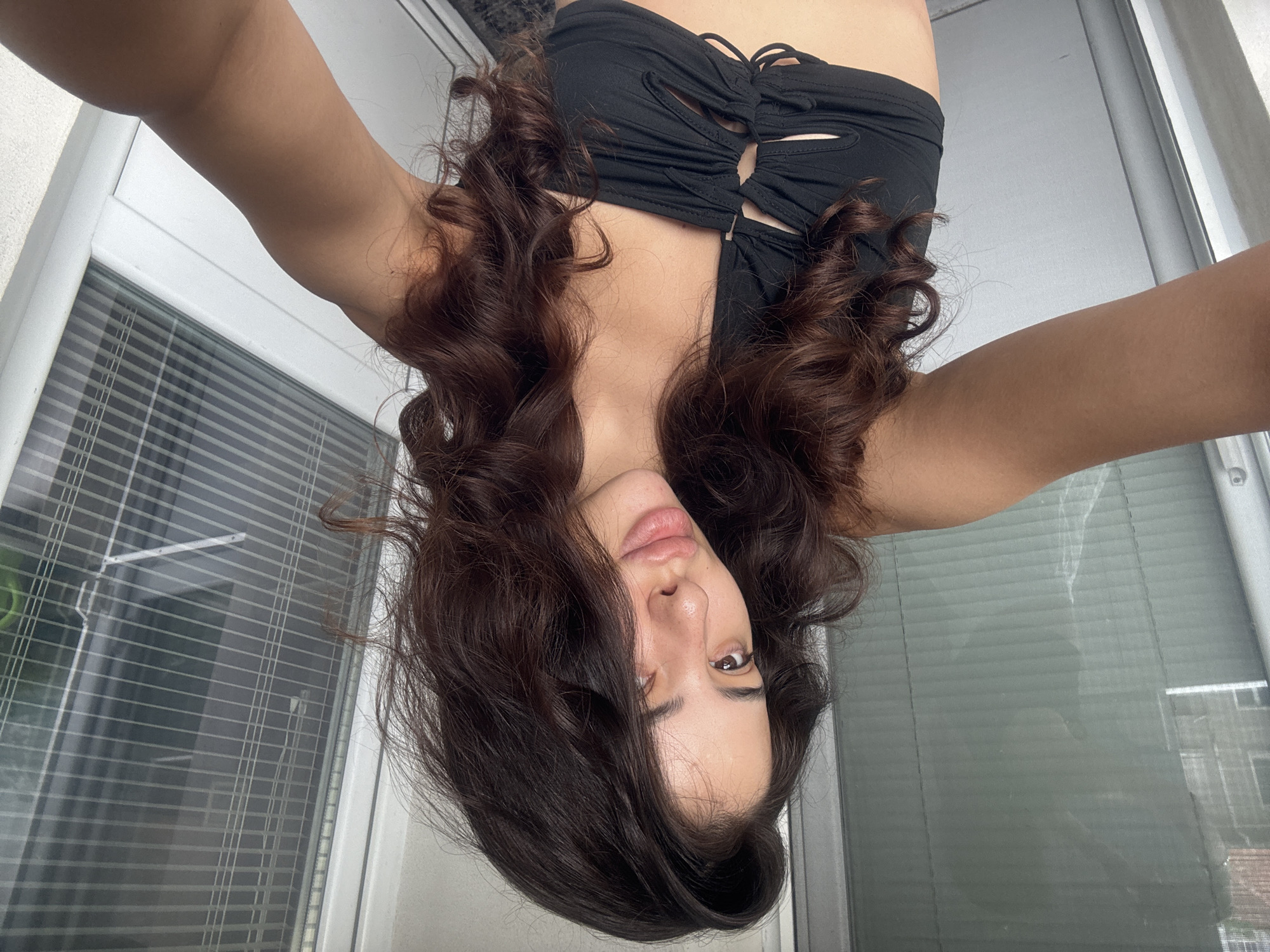 The media file shows a young woman taking a selfie. She is upside down, with her face in the foreground and her arms and torso extending upwards. She has long, dark, curly hair and is wearing a black, strappy bandeau top. Her expression is neutral, with her eyes open and looking towards the camera. The background appears to be an indoor setting, possibly a room with a window that has horizontal blinds. The light suggests it is daytime. The woman's skin is smooth and unblemished. Her arms are raised, and her hands are out of frame, implying she is holding a phone to take the picture. The scene is well-lit, and the focus is clearly on the woman. There is no visible text. The overall impression is a casual, personal photograph.