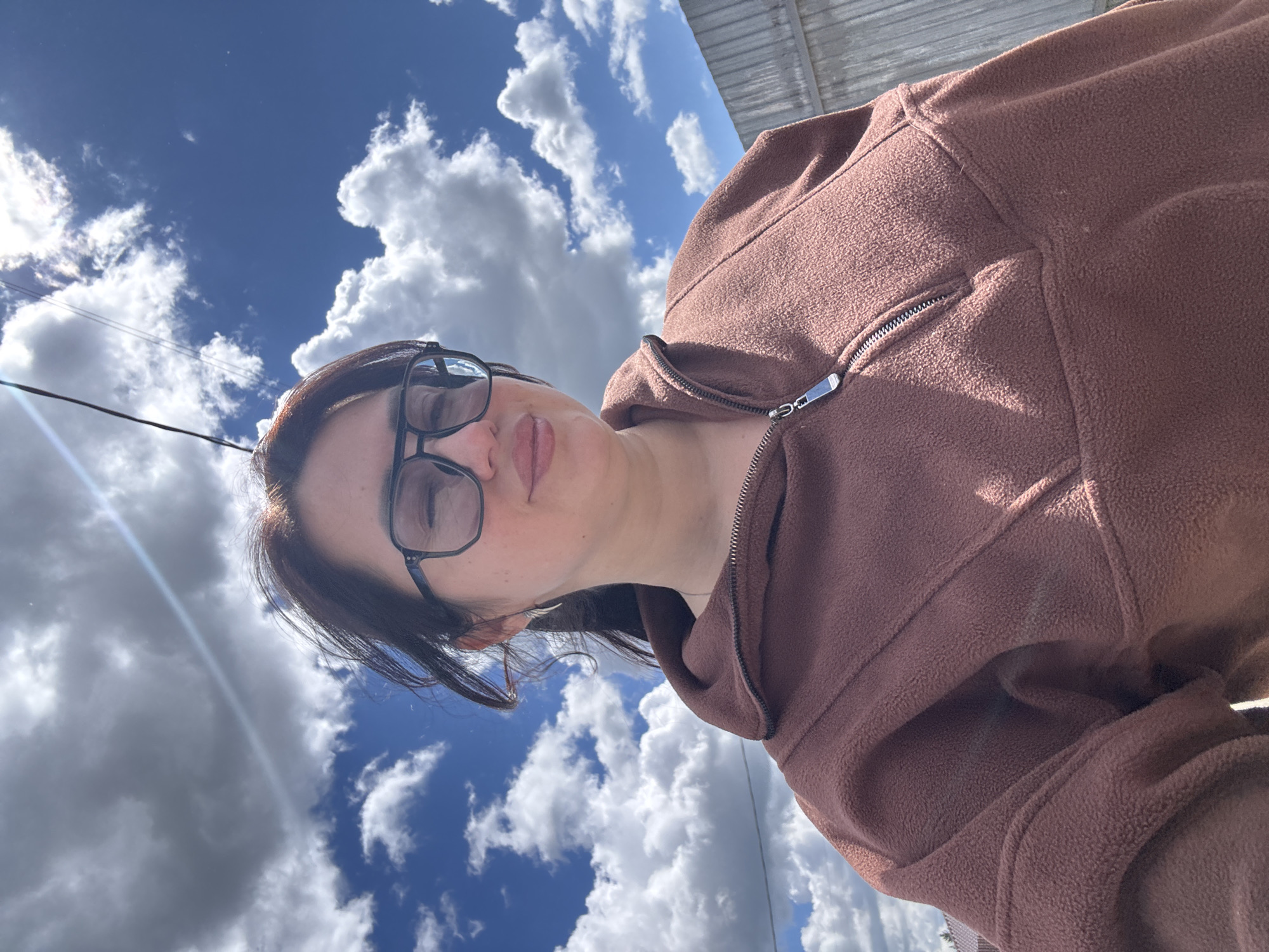 The image shows a selfie of a woman wearing glasses and a brown hooded jacket, taken against a backdrop of a blue sky with white clouds. The location is unknown, as is the city. The woman is looking at the camera with a neutral expression. A power line is visible in the upper left corner of the frame. The background includes what appears to be the edge of a building. The image is slightly tilted.