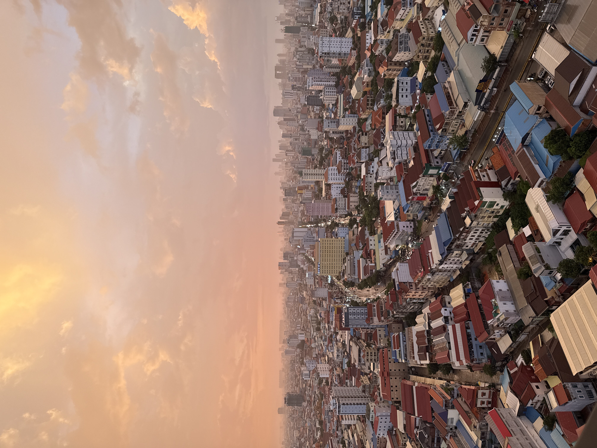 The image captures an aerial view of Khan Mean Chey, Cambodia, under a dramatic sky filled with orange and pink clouds, indicative of either sunrise or sunset. The urban landscape is densely packed with a mix of low-rise buildings with red and blue roofs, as well as taller, modern high-rises dominating the skyline in the distance. Streets are visible, some lined with vehicles, suggesting active traffic within the city. The atmosphere appears somewhat hazy, common in urban environments, especially towards the horizon where the city fades into the distance. There are no distinct individuals or specific actions clearly identifiable from this high vantage point, but the scene clearly depicts a bustling urban environment at dusk or dawn.