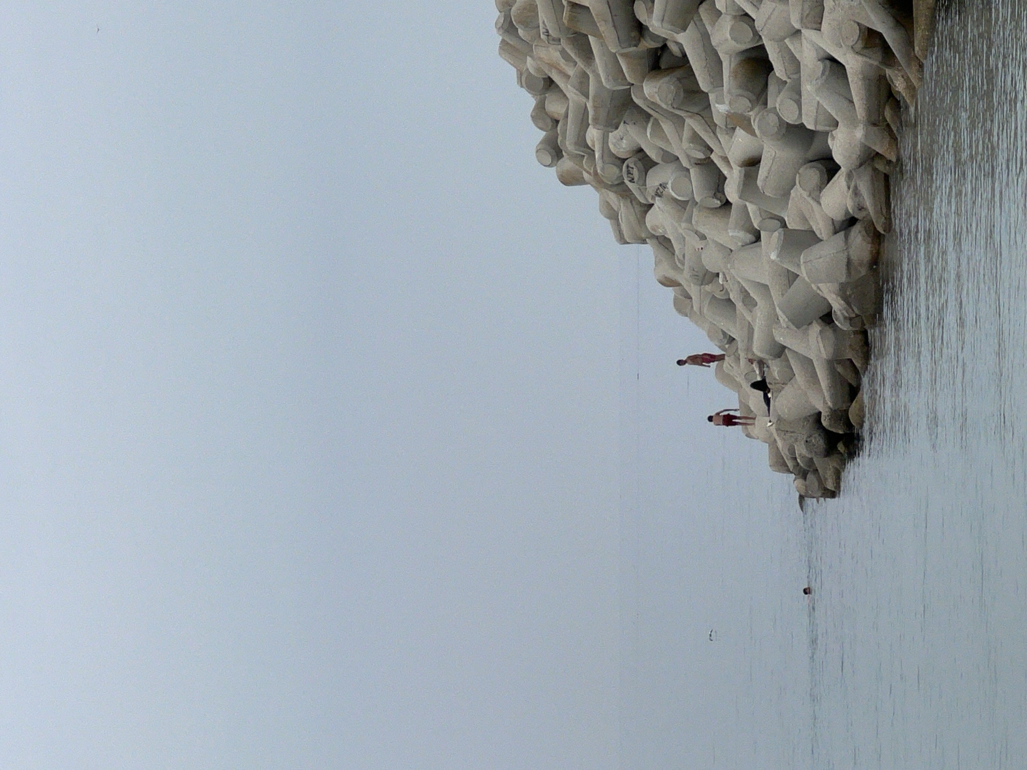 A serene coastal scene unfolds under an overcast sky, featuring a substantial concrete breakwater alongside tranquil waters. The breakwater, constructed from numerous interlocking, light grey and beige-toned concrete blocks (likely tetrapods or accropodes), dominates the right side of the image, extending diagonally downwards towards the middle.
Three individuals are present. Two are positioned on the concrete breakwater; one person, wearing reddish swimwear, stands upright on the blocks, appearing to gaze at the water or prepare for a swim. Further along the breakwater, another individual is partially visible, seemingly sitting or crouching among the blocks. The head of a third person is visible in the calm, light blue-grey water, swimming or floating further away from the structures. A small, dark spherical object, possibly a buoy or ball, floats in the water closer to the foreground.
The sky, which occupies the left side of the frame, is a uniform, soft light blue-grey, indicating overcast conditions with diffused lighting. The water surface is calm, gently reflecting the sky. Close examination of the concrete blocks reveals faint, dark markings or text, such as "N.T." and "N.Z.0." or "N.2.0.", which are likely identification or manufacturing codes. The scene captures a quiet moment of leisure and interaction with the sea in M'Diq, Morocco.