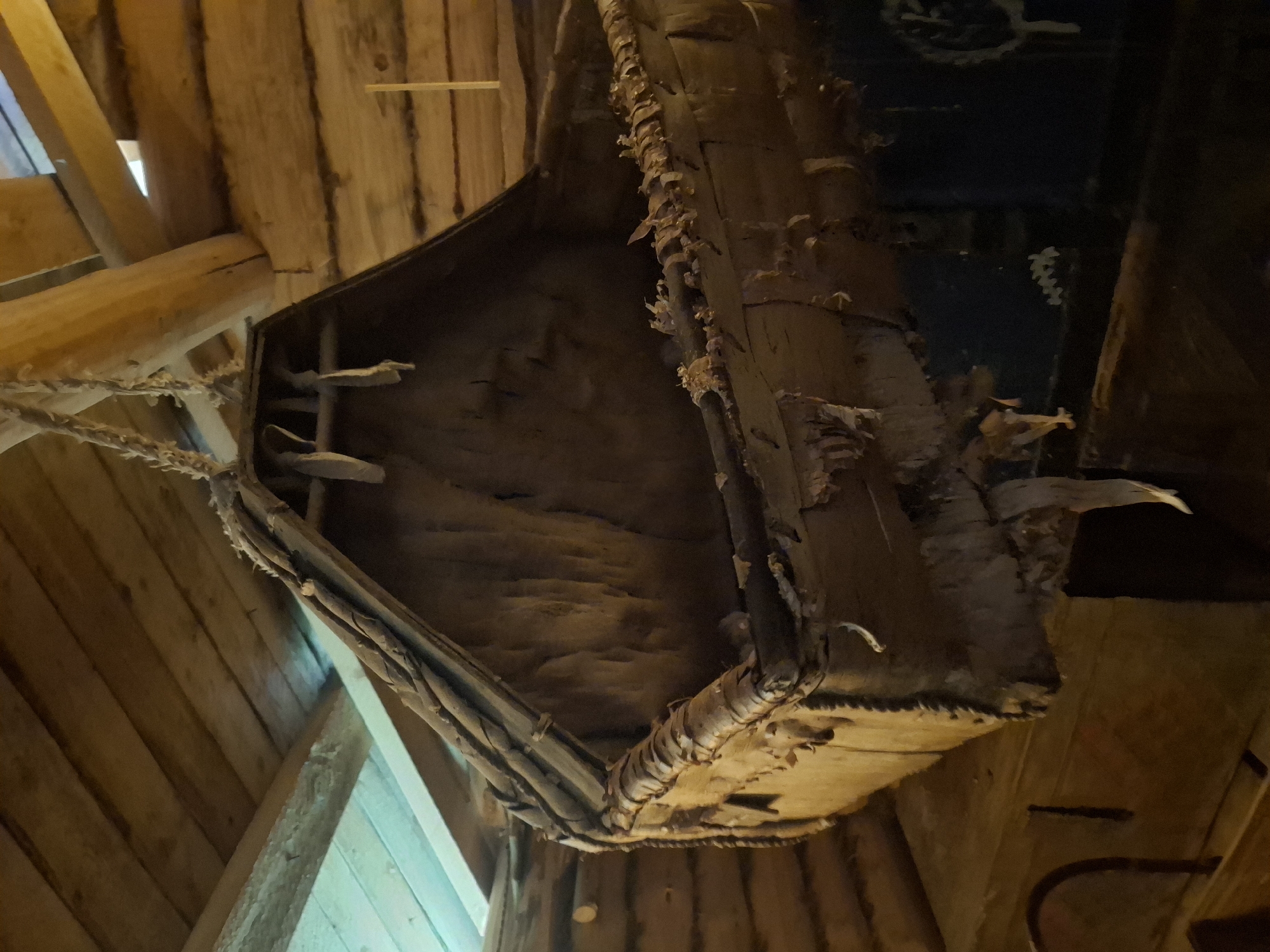 The image shows a hanging birch bark container inside a wooden structure, likely a building. The container is suspended by ropes and appears to be constructed of birch bark and wooden supports. The surrounding structure is made of unfinished wood planks and beams. The location is unspecified, but the building style suggests a historical or rural setting.