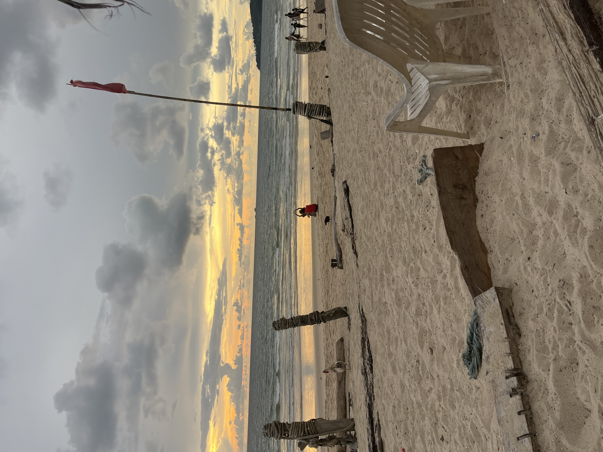 This media file depicts a beach scene at dusk in Choeng Thale, Thailand. The horizon is filled with a dramatic sunset, with vibrant yellow and orange hues piercing through overcast, gray clouds. The sky transitions from dark gray at the top to a bright, luminous band of color near the horizon.
On the sandy beach, several objects and elements are visible. A red flag flies from a tall, slender pole in the foreground. To the right, a white plastic beach chair is positioned on the sand. Several weathered wooden posts, some wrapped with rope, are planted in the sand, suggesting a possible fence or demarcation. A person in a red garment is visible kneeling by the water's edge. Further in the distance, a group of people are walking along the shoreline.
The scene evokes a sense of tranquility and the end of a day. The calm sea gently laps at the shore. The overall atmosphere suggests a peaceful, natural setting, possibly a tourist beach. No visible text is present in the image.
