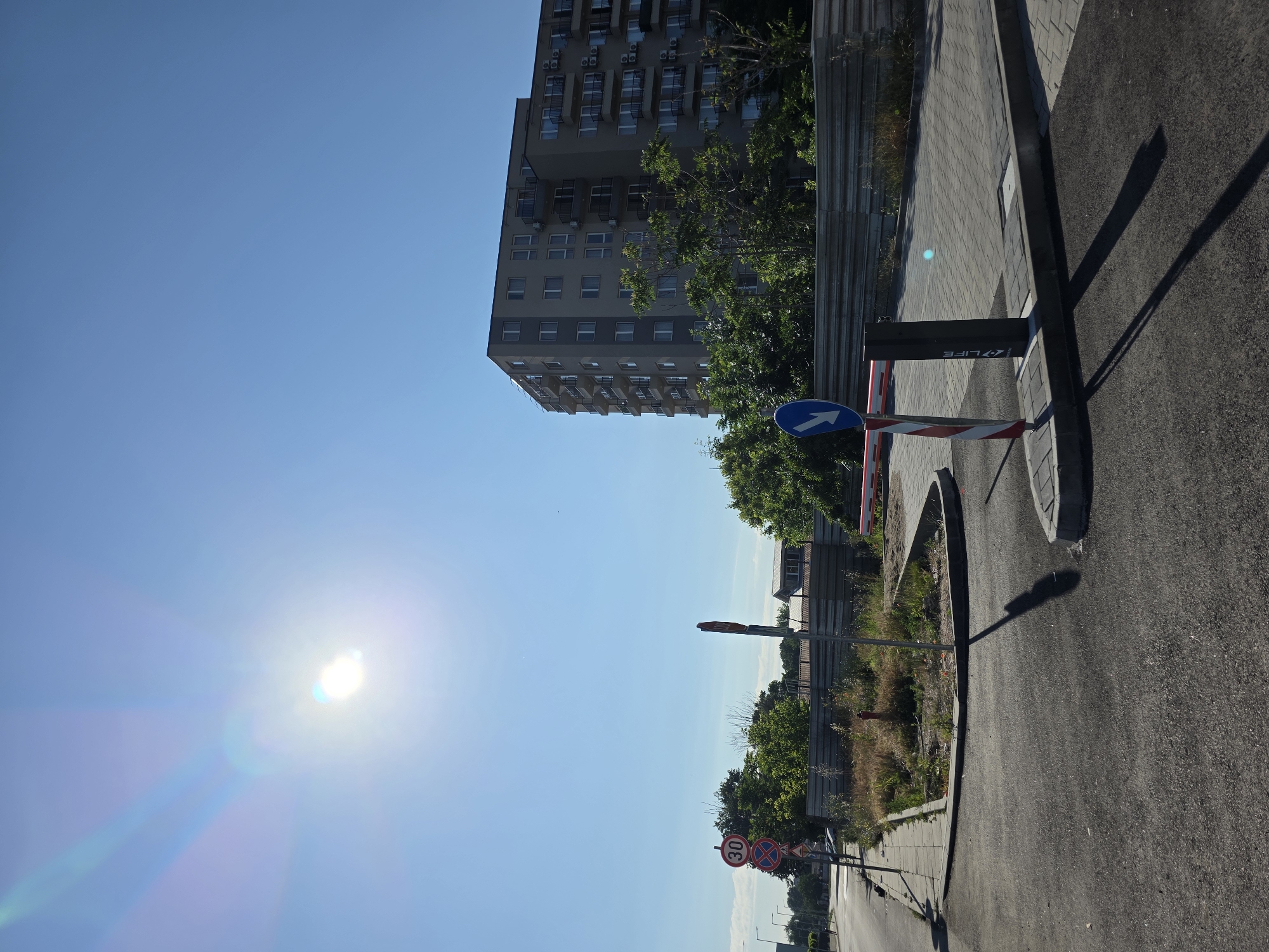 A bright, sunny day with a clear blue sky is depicted in an outdoor scene. The sun, positioned in the upper left quadrant, creates a lens flare effect. In the middle ground, a multi-story residential building with many balconies and windows stands tall, partially obscured by lush green trees. To the left of the building, a street is visible, lined with streetlights and the edge of a sidewalk. Several traffic signs are present, including a circular sign with the number "30" inside and another diamond-shaped sign with a red cross and a red border, indicating traffic regulations.
In the foreground, a paved area with cobblestone accents leads to an automated barrier gate, likely for vehicle access control, with a red and white striped pole and a black electronic unit. The ground is asphalt, showing tire marks and shadows cast by the objects and structures. A small patch of overgrown weeds and dry grass with a red fire hydrant is situated between the sidewalk and the barrier gate. The overall impression is a scene in a developed urban area, possibly an entrance to a residential complex or a controlled access point. The mention of Plovdiv, Bulgaria, suggests this location is within that city.