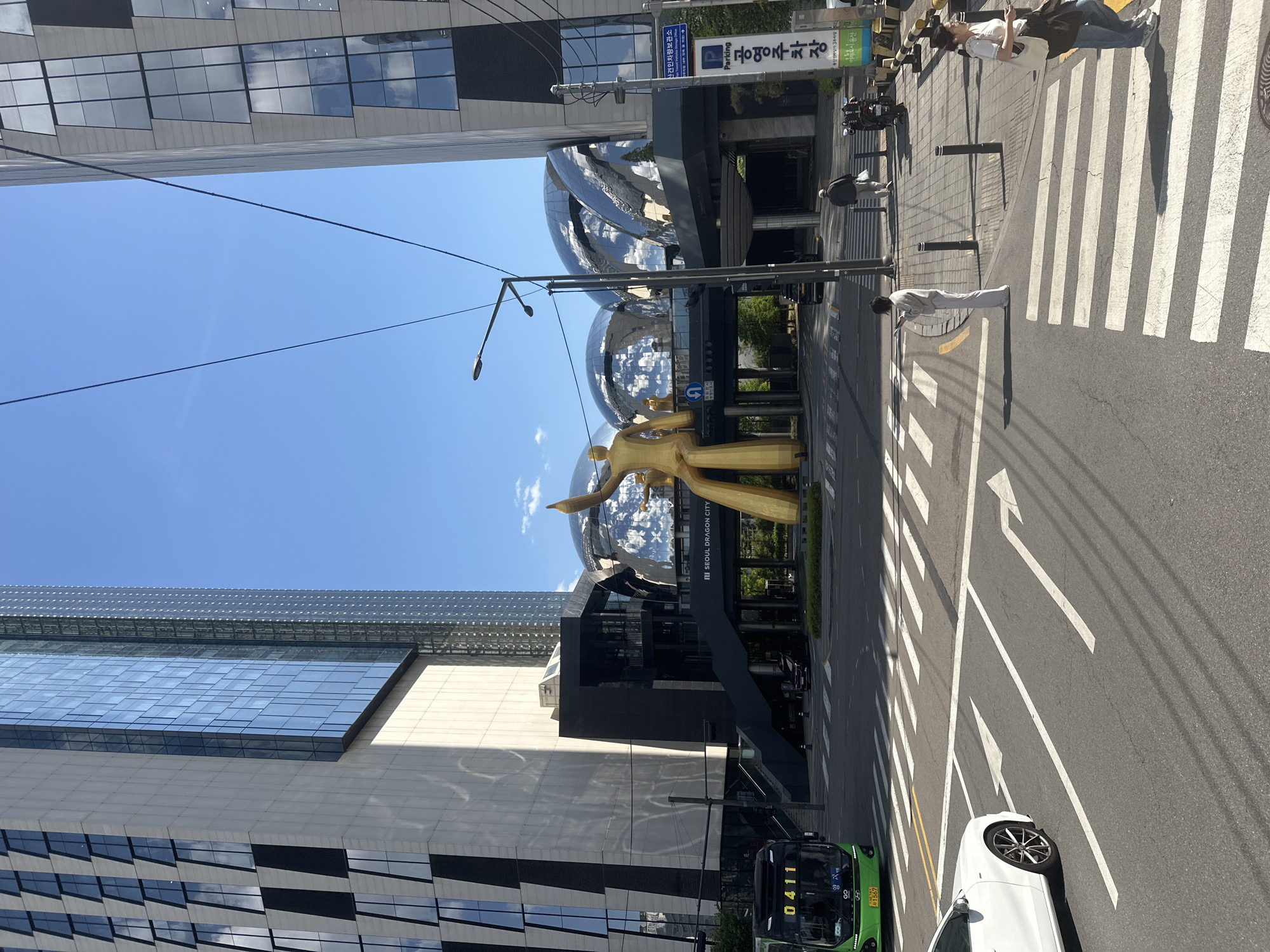 Here is a detailed description of the image:
This is an eye-level outdoor shot, rotated at ninety degrees, that features a street scene in Seoul, South Korea. Prominent in the scene is a golden statue depicting two human figures in front of a building complex. This complex has a modern architectural design with a gray and black facade. Above the statue, two large, spherical, reflective architectural features catch the sunlight. Inscription on the building reads "SEOUL DRAGON CITY".
To the right of the complex, there is a crosswalk with pedestrians, including one crossing with a long shadow indicating the direction of the sun. A green bus and a white car are visible on the road. The blue sky provides a backdrop to the building structures on either side. Street signs written in Korean are also visible.
