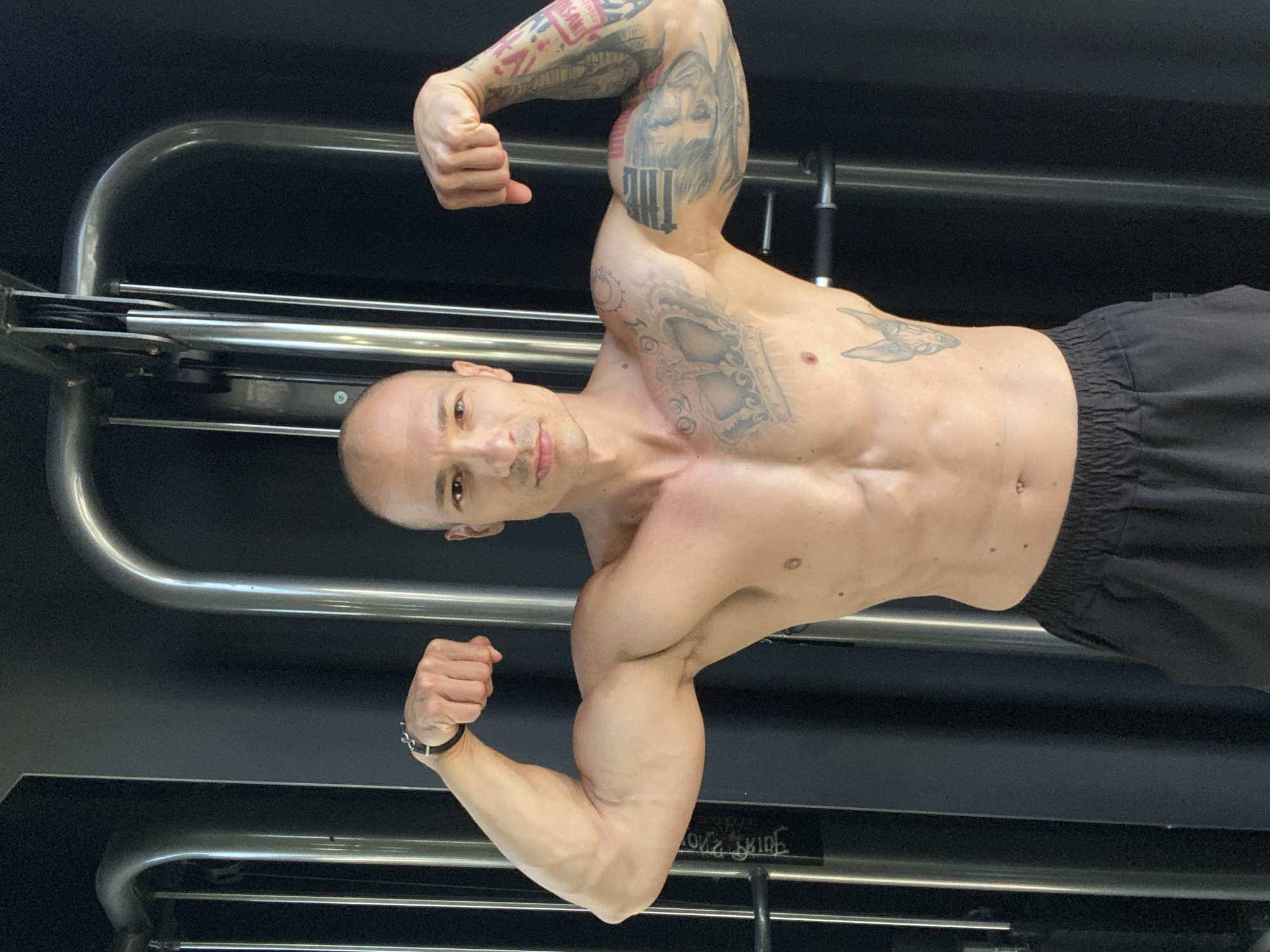 A bald, muscular man with tattoos on his chest and arms poses with his biceps flexed in a gym. He is wearing black shorts. The background consists of gym equipment. The image was taken in Pleven, Bulgaria.