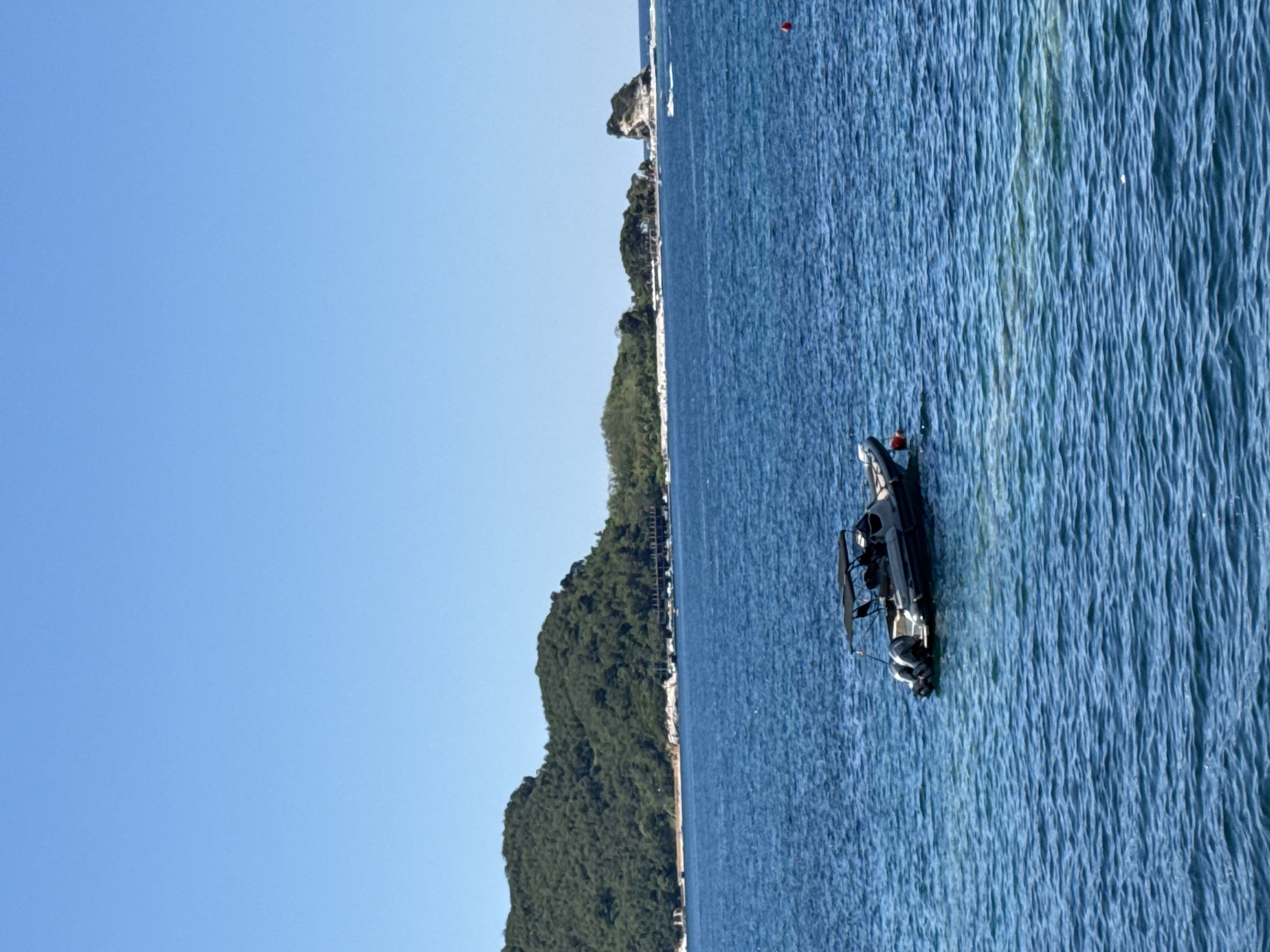 The image shows a serene coastal scene in Budva, Montenegro. In the foreground, a small, gray rigid inflatable boat with a black canopy is anchored on the calm, blue water. The boat has two outboard motors. In the middle ground, a lush green, tree-covered hill rises from the coastline, with a few buildings visible along its base. Further in the distance, small, rocky islands dot the sea, one with a distinctive rock formation. The sky is a clear, bright blue. The overall impression is one of tranquility and natural beauty.