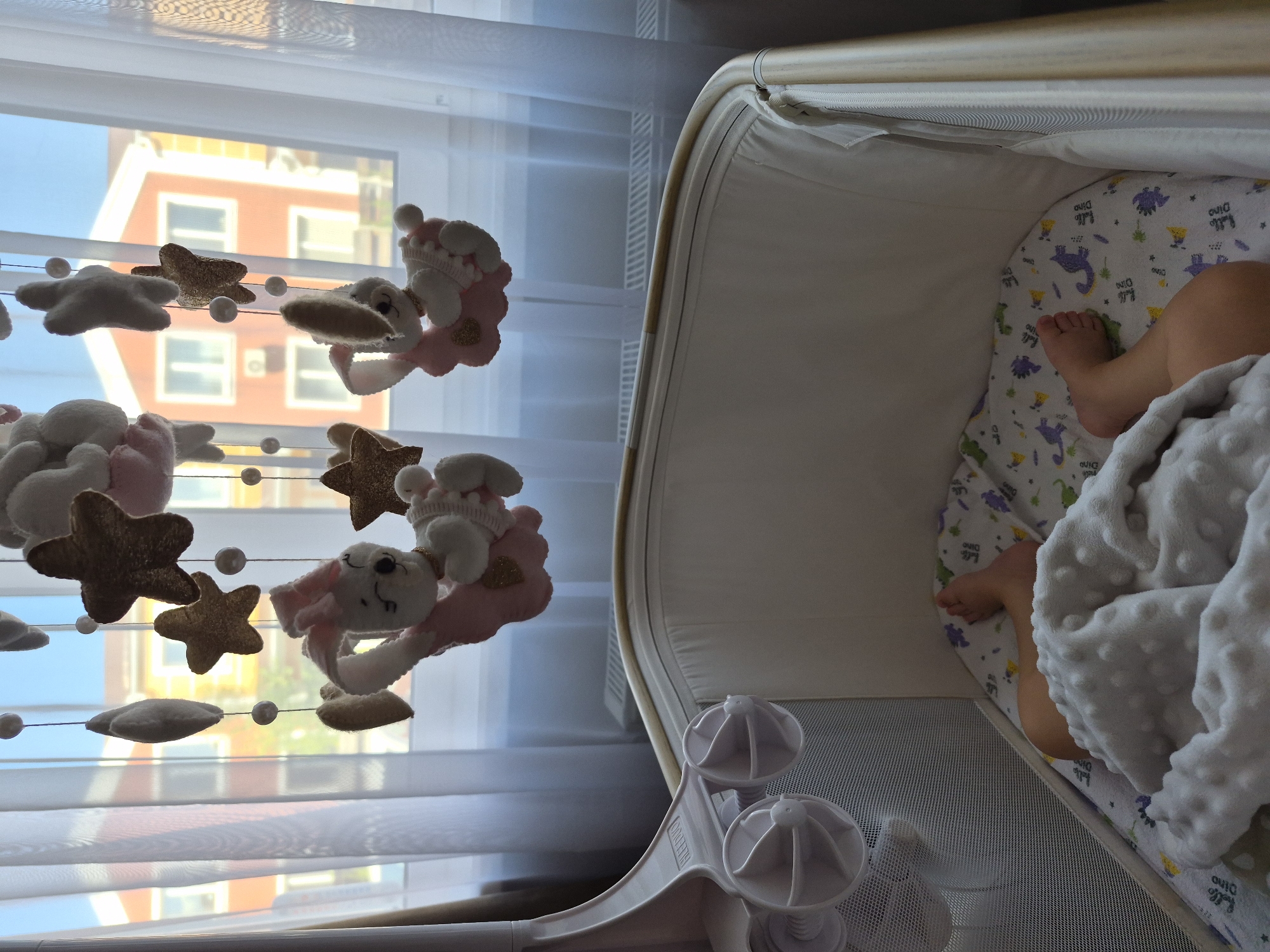 In an indoor scene, a baby's legs are visible extending from under a white, textured blanket in a crib. The crib has a white mattress with a colorful, patterned sheet featuring dinosaurs and the words "Hello Dino." A mobile with plush bunnies and stars hangs above the crib. Through a sheer white curtain in the background, two houses with orange and yellow facades and white-trimmed windows are visible, suggesting an urban or suburban setting. Sunlight streams in, illuminating the room.