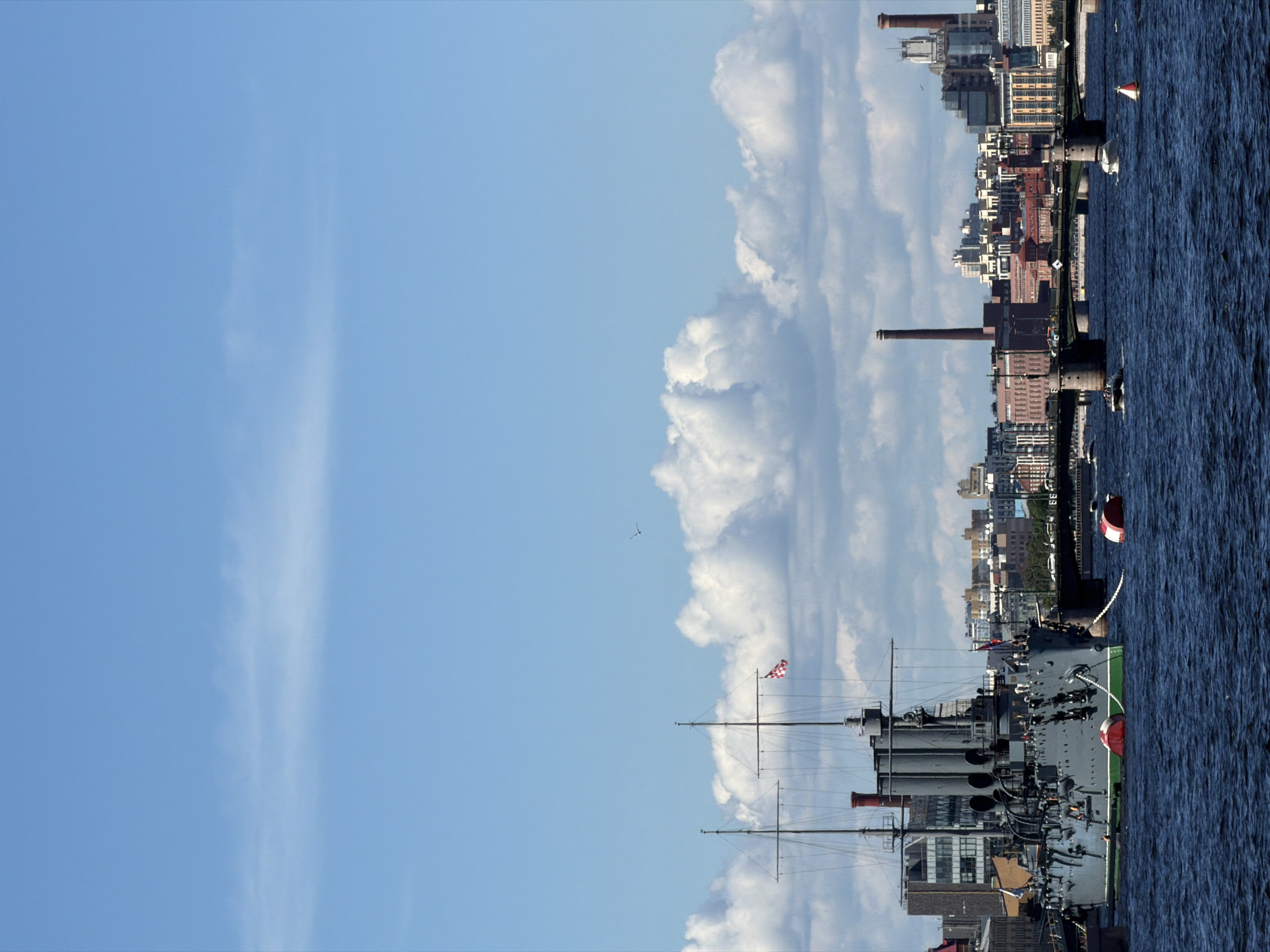 The image displays a wide, daytime view of the Neva River in Saint Petersburg, Russia, under a bright blue sky with scattered white cumulus clouds. In the foreground, the Cruiser Aurora is anchored on the left, presenting a grey hull with a green waterline, two masts, and three prominent funnels. A red and white checkered flag is flown from its aft mast. Two large red and white cylindrical buoys are moored near the ship's bow, connected by anchor chains. The river's surface is dark blue and choppy. Several smaller motorboats, including a speedboat and a white cabin cruiser, navigate the water, along with a solitary red and white conical buoy.
In the midground, a multi-arched bridge with stone piers and a dark metal superstructure spans the river, carrying vehicular traffic visible as small, moving forms. Beyond the bridge, the cityscape of Saint Petersburg forms a dense backdrop, comprising a variety of multi-story buildings of differing architectural styles and ages. Two tall, reddish-brown brick industrial chimneys rise distinctly above the skyline on the right. High in the sky, a faint, linear cloud streak or contrail is visible across the upper portion of the frame, and a small, dark bird is captured in flight above the city's horizon line. Small human figures are discernible on the deck of the Aurora and within some vehicles on the bridge.