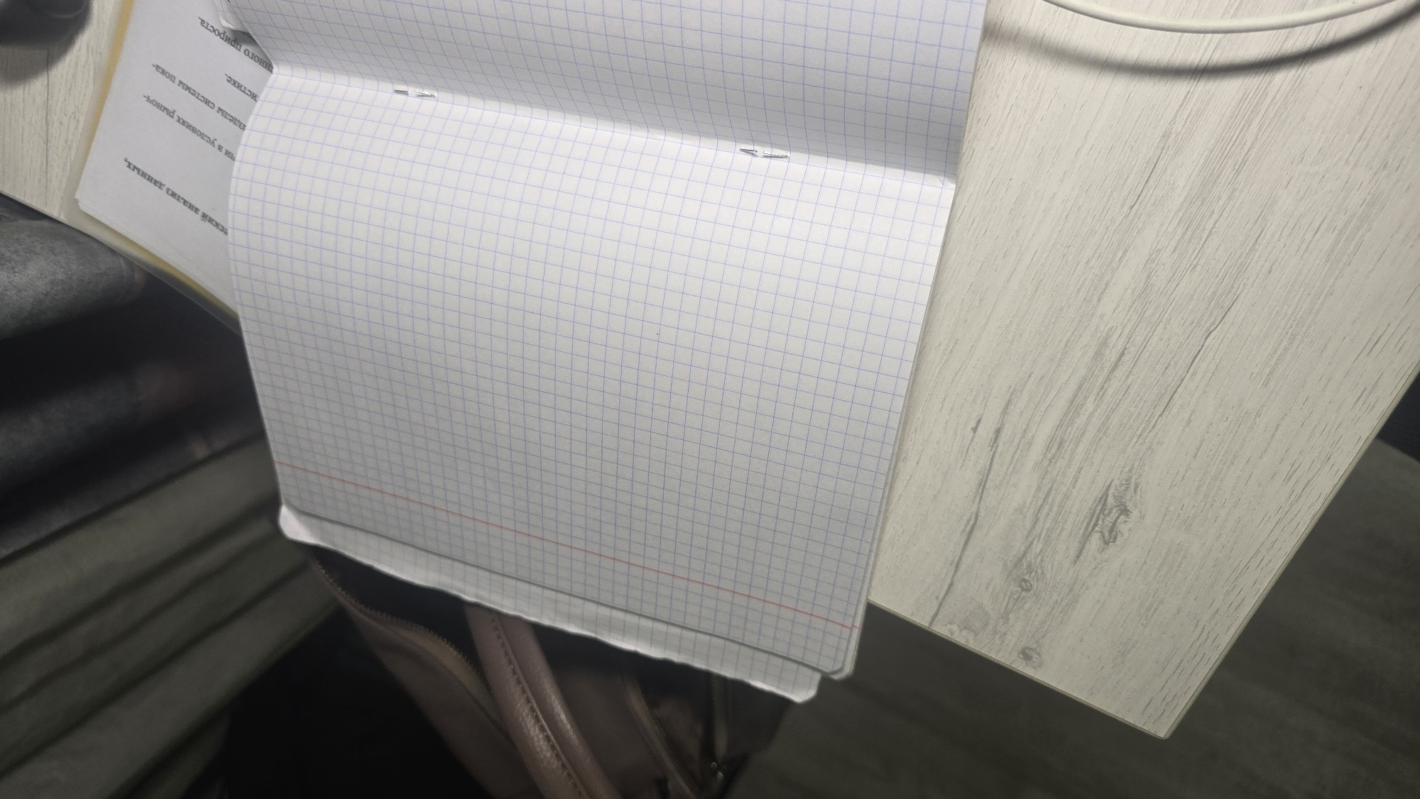 This media file is a close-up, high-angle shot of an open notebook with graph paper, resting on a light-colored, wood-grain textured surface. The notebook is positioned at an angle, with the right side of the page facing upwards. A portion of a white cable is visible at the top right of the image, suggesting it might be connected to a device or charging. The background is blurred, showing dark, indistinct shapes that could be furniture or other objects.
The notebook pages have faint blue grid lines and a red horizontal line near the bottom of the visible page. There are some staples holding the pages together, visible on the left side of the open notebook.
There is visible text on the left-hand page of the notebook, written in a dark font. The text appears to be in Russian and includes phrases like "еский анализ данных" (possibly referring to data analysis), "в условиях рыноч" (in market conditions), "системы пока-" (system shows), and "ного прироста" (growth). The text is oriented such that it reads upwards from the bottom of the page.
The lighting suggests an indoor setting, possibly an office or study area, with artificial light. The overall impression is one of a workspace with a focus on a notebook containing written information. No people are visible in the frame. The scene is static and appears to be a still photograph rather than a video. The texture of the table and the paper are prominent details. The context of Ukhta, Russia, is provided but not visually evident within the image itself.