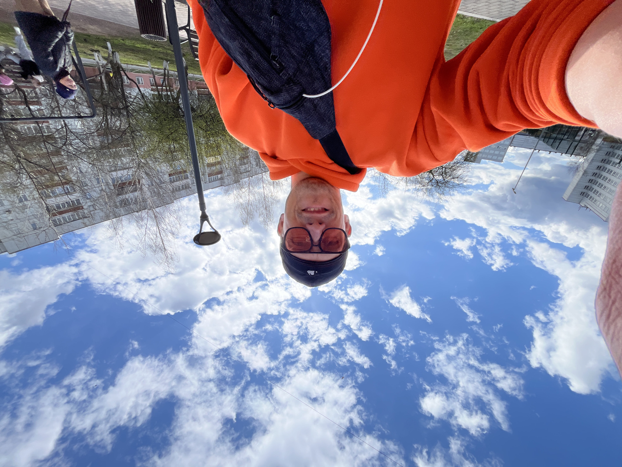A low-angle selfie captures a smiling man looking upwards at the camera, set against a backdrop of a bright blue sky with white clouds and an urban environment. The man is wearing a vibrant orange hooded sweatshirt, a dark fanny pack across his chest, dark-rimmed glasses, and a dark baseball cap. A white earbud wire is visible near his left shoulder. His face displays a pleasant, content smile with visible stubble.
The upper half of the image is dominated by a clear blue sky filled with numerous white, fluffy cumulus clouds, indicating a sunny day. In the midground and background, an urban or suburban landscape unfolds. A tall, slender dark street lamp pole extends upwards on the left side of the frame. Beyond it, several multi-story buildings, appearing to be residential or office structures with many windows, are visible. Some trees have bare branches, while others show early signs of light green foliage, suggesting it is early spring. A paved path or sidewalk runs along the bottom, with patches of green grass. Faint figures of other people, including what looks like a woman and a child, can be seen walking on the distant path.
The scene depicts an outdoor moment during the daytime, under bright and pleasant weather conditions. The man's expression conveys happiness or contentment. The setting aligns with an urban area, consistent with Chaikovsky, Russia. No discernible text is visible within the image.