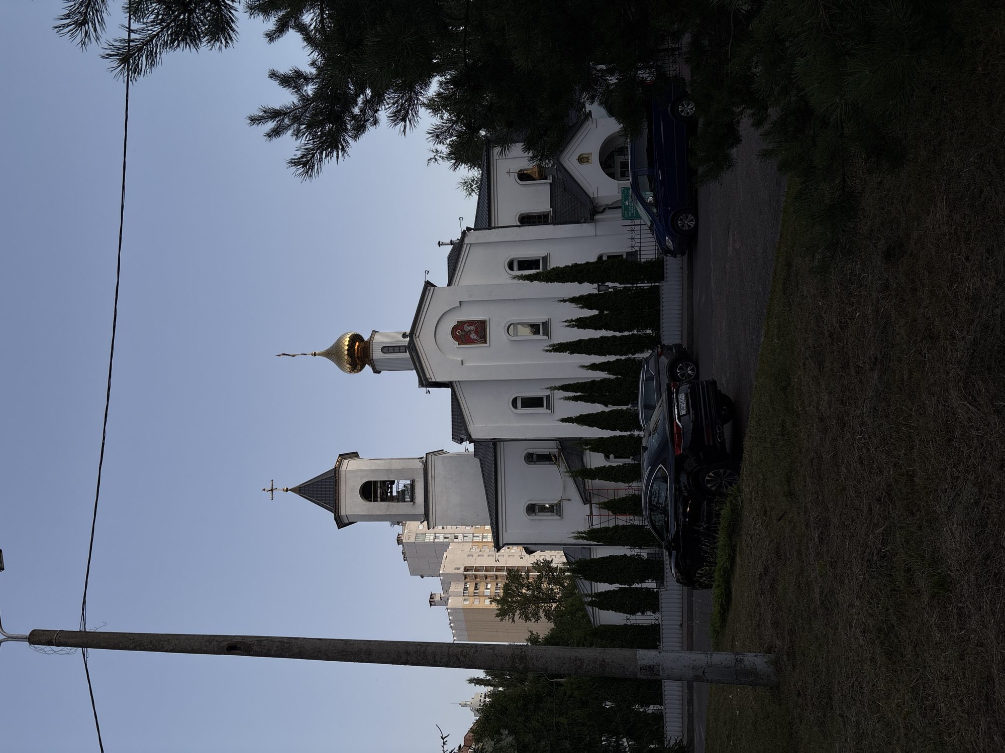 This image depicts the Church of the Holy Trinity in Vitsebsk, Belarus. The church is a white building with a dark tiled roof, featuring a bell tower with a cross and a golden dome topped with a cross. In front of the church, a black car is parked on the street, and a blue van is parked further down. Tall evergreen trees line the front of the church, and a metal fence separates the church property from the street. In the background, several tall apartment buildings are visible. The sky is clear and blue. The foreground shows a grassy area with a concrete lamppost on the left side of the frame.