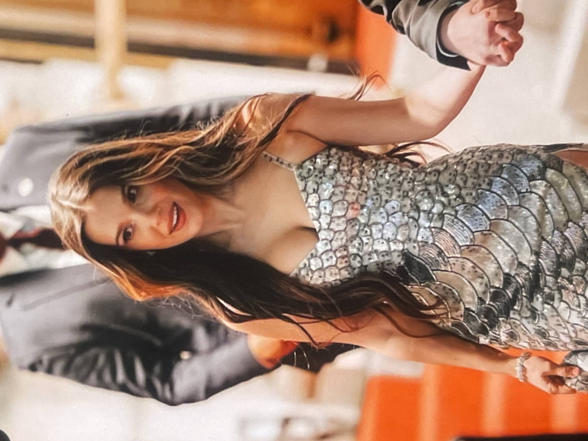 This rotated portrait-oriented photograph captures a woman smiling at the camera while holding hands with another person, whose body is mostly out of frame. The woman has long, wavy dark brown hair and is wearing a striking sleeveless dress adorned with numerous silver or iridescent sequins arranged in a scale-like pattern. Her smile suggests happiness or engagement. On her visible right hand, she wears a ring or bracelet.
The hand she is holding belongs to another individual, partially visible in the upper right portion of the frame, wearing what appears to be a dark or grey long-sleeved shirt or jacket. The grip suggests a firm, connected hold.
The background is softly blurred, displaying warm, orange-toned elements mixed with darker, indistinct shapes, indicating an indoor setting. There are no specific architectural or environmental cues within the image itself to confirm the exact location beyond the provided context of Poltava, Ukraine. The lighting appears to be artificial, suggesting it could be day or night indoors. There is no visible text in the image. The overall impression is a joyful and intimate moment shared between the subjects.