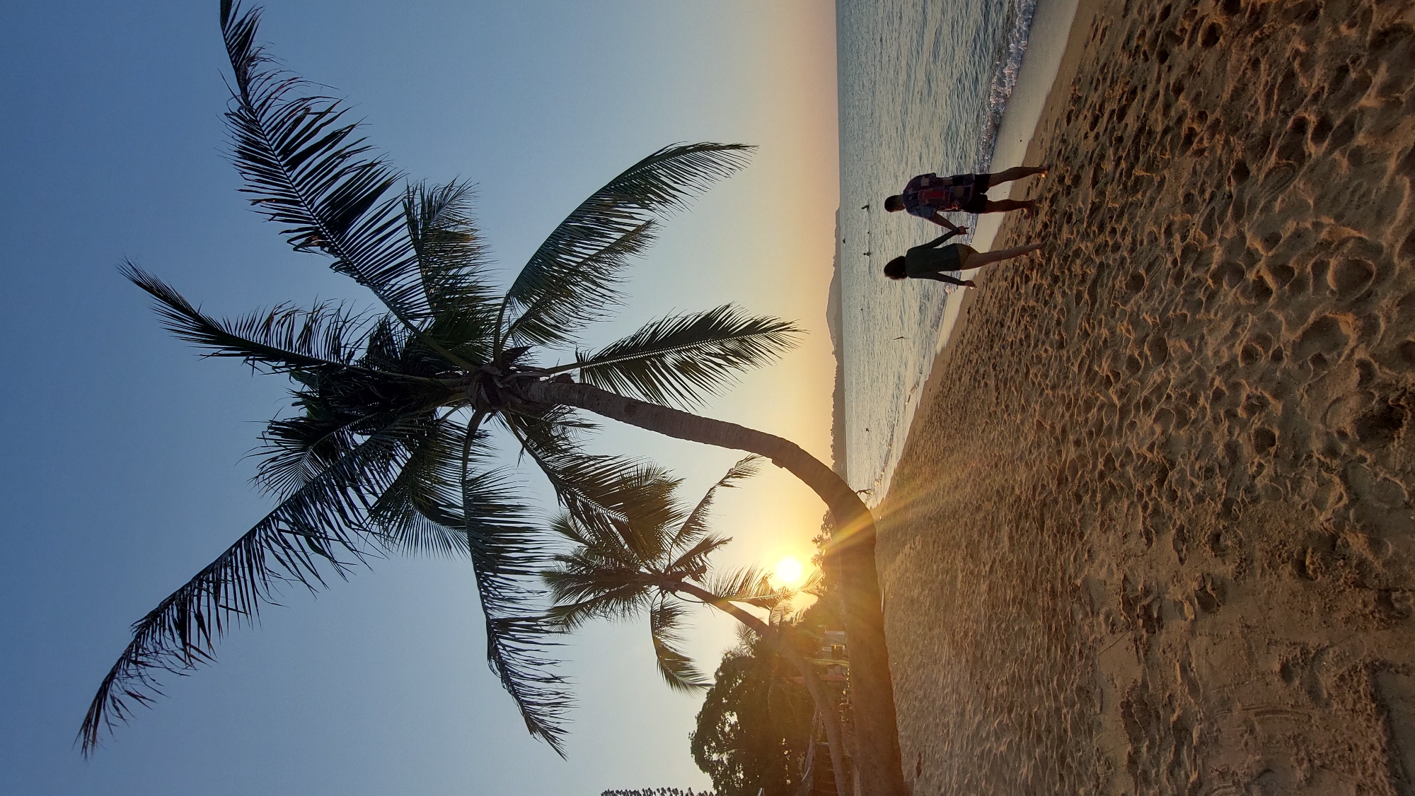 A beach scene at sunset shows a palm tree leaning to the left against a clear blue sky. Two people walk on the sandy beach near the water's edge. The setting sun peeks through the trees. The beach is marked with footprints. The location is Baan Mae Nam, Thailand.