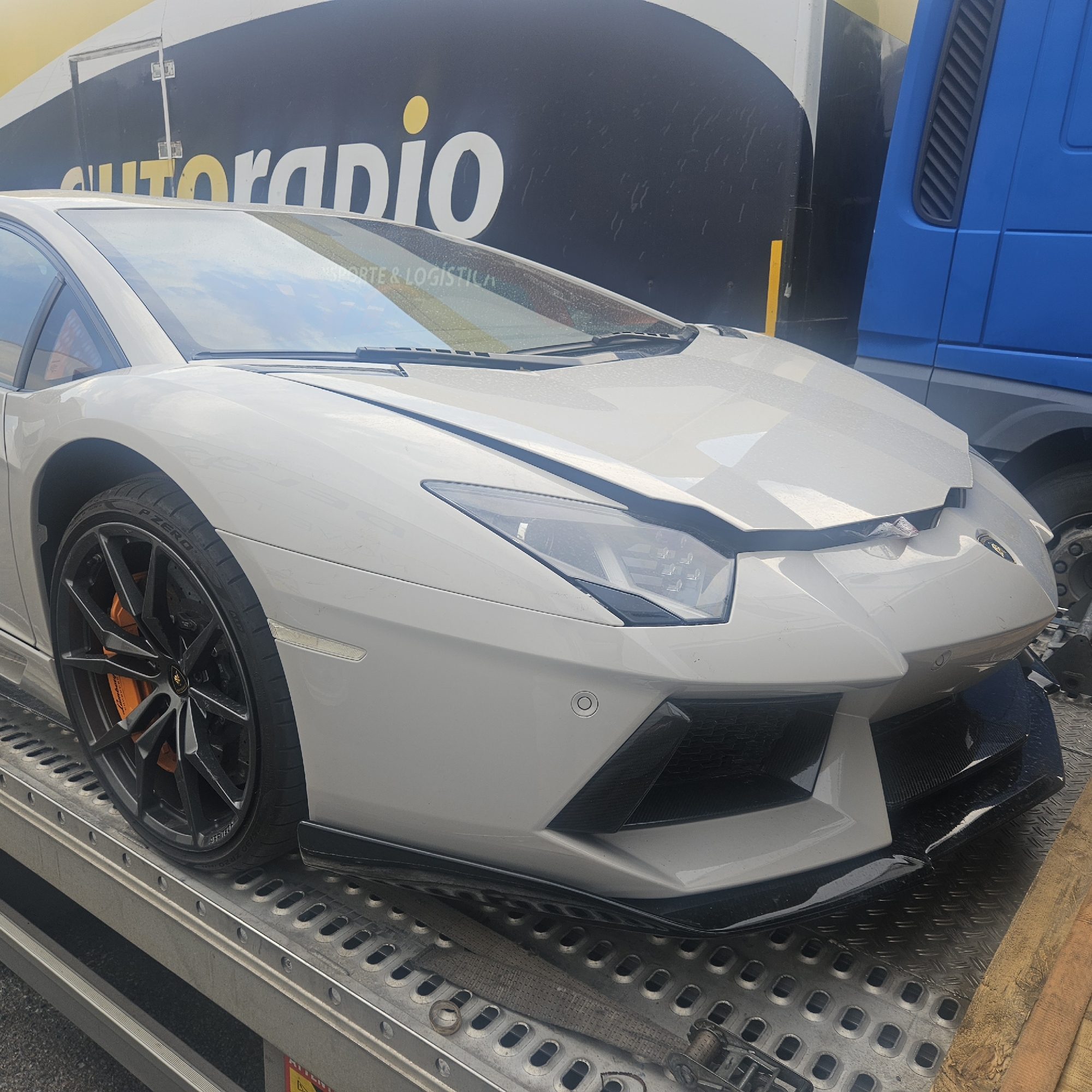 A silver Lamborghini Aventador is loaded onto a car transport trailer. The front of the car shows damage to the hood and bumper. The car is positioned on the lower deck of the trailer, which is grey and metallic with circular cutouts. In the background, a large yellow and black truck with the logo "AUTORADIO" is visible, along with the side of a blue truck. The scene appears to be in La Jonquera, Spain, as indicated by the context of the location. There are no people clearly visible in the image. The environment suggests an outdoor setting, likely a logistics or transport hub.