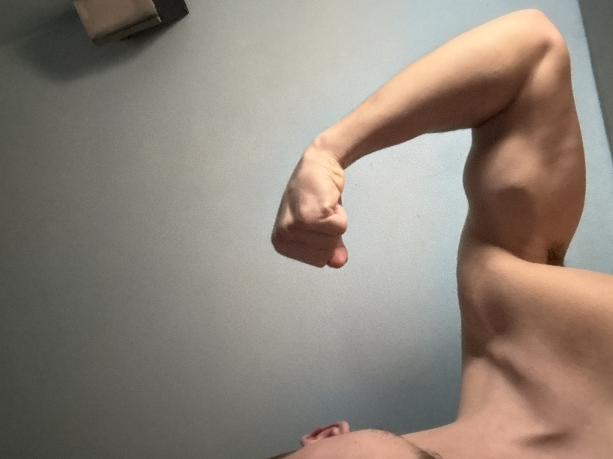 A close-up, low-angle shot depicts a muscular arm flexing. The arm is bare, and the hand is clenched into a fist. Parts of the neck, upper chest, and ear are visible at the bottom of the frame. The background is a plain gray wall with a small object attached near the top left. There is no indication of the location being Buhovo, Bulgaria.