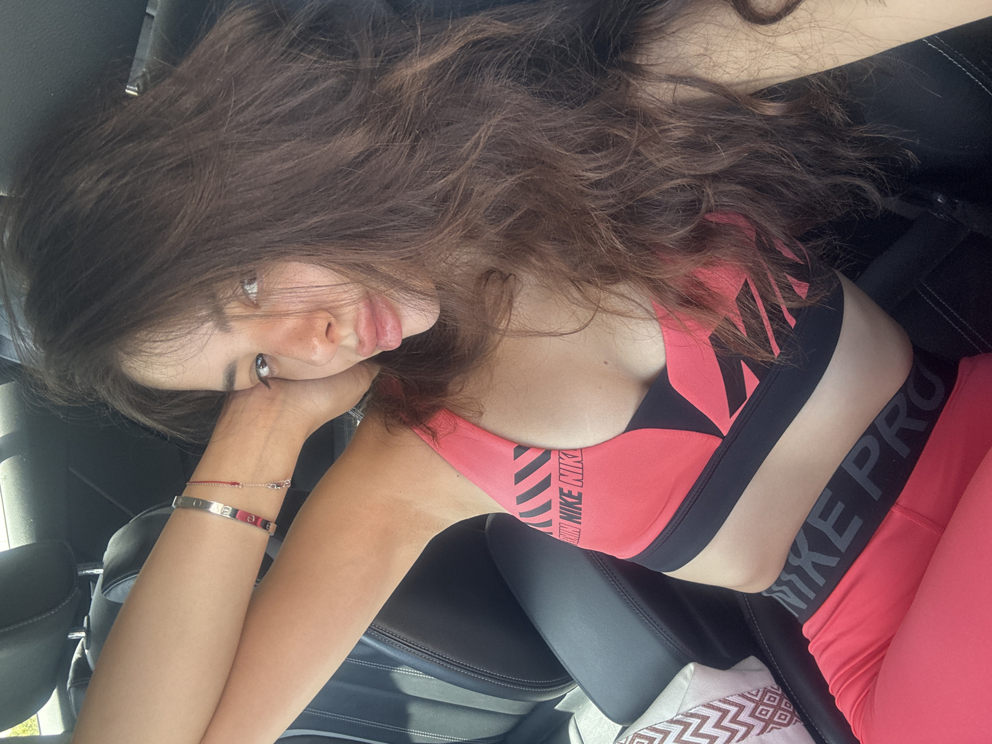 A young woman with long, dark, wavy hair is sitting in the driver's seat of a car, looking at the camera. She is wearing a pink sports bra with black accents and the "Nike Pro" logo, and matching pink leggings. Her right arm is resting behind her head, and her left arm is bent at the elbow, with her hand near her head. She is wearing a silver bracelet on her left wrist. The car interior is visible, with black leather seats. The scene appears to be outdoors, with sunlight streaming into the car. The image does not provide enough information to determine the specific location within the city of Plovdiv, Bulgaria.