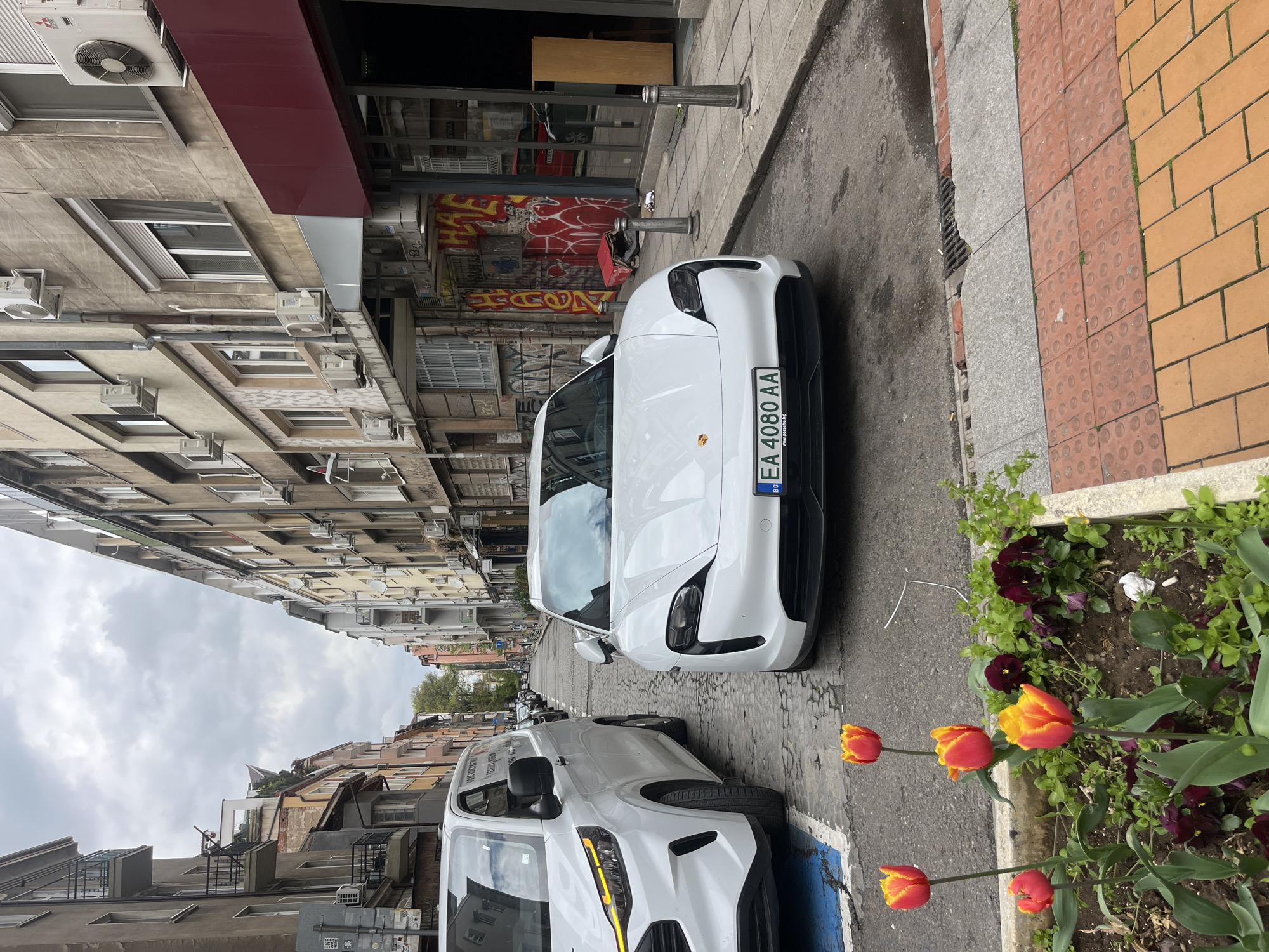 The image shows a street scene in Sofia, Bulgaria. A white Porsche Taycan with the license plate "EA 4080 AA" is parked on the right side of the street, close to a planter box with red and yellow tulips, plus dark purple pansies. A white van is partially visible parked behind the Porsche, also appearing to be parked on the street. The street itself is narrow, paved with cobblestones, and lined with buildings. The buildings are multi-story, with a mix of architectural styles. Graffiti is visible on the building across the street from the Porsche. The sky is overcast.