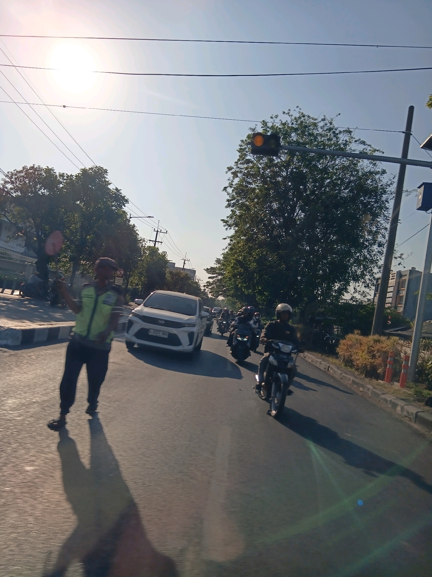 In Surabaya, Indonesia, a traffic officer in a yellow vest and dark pants directs traffic on a sunny day. The officer is holding a red traffic sign in their left hand. The scene is a street with multiple lanes of traffic. Several motorcycles and a white car are visible. A traffic light, with its amber signal illuminated, hangs overhead. Trees line the side of the road, and buildings are visible in the background. The sun is bright, casting long shadows on the asphalt.