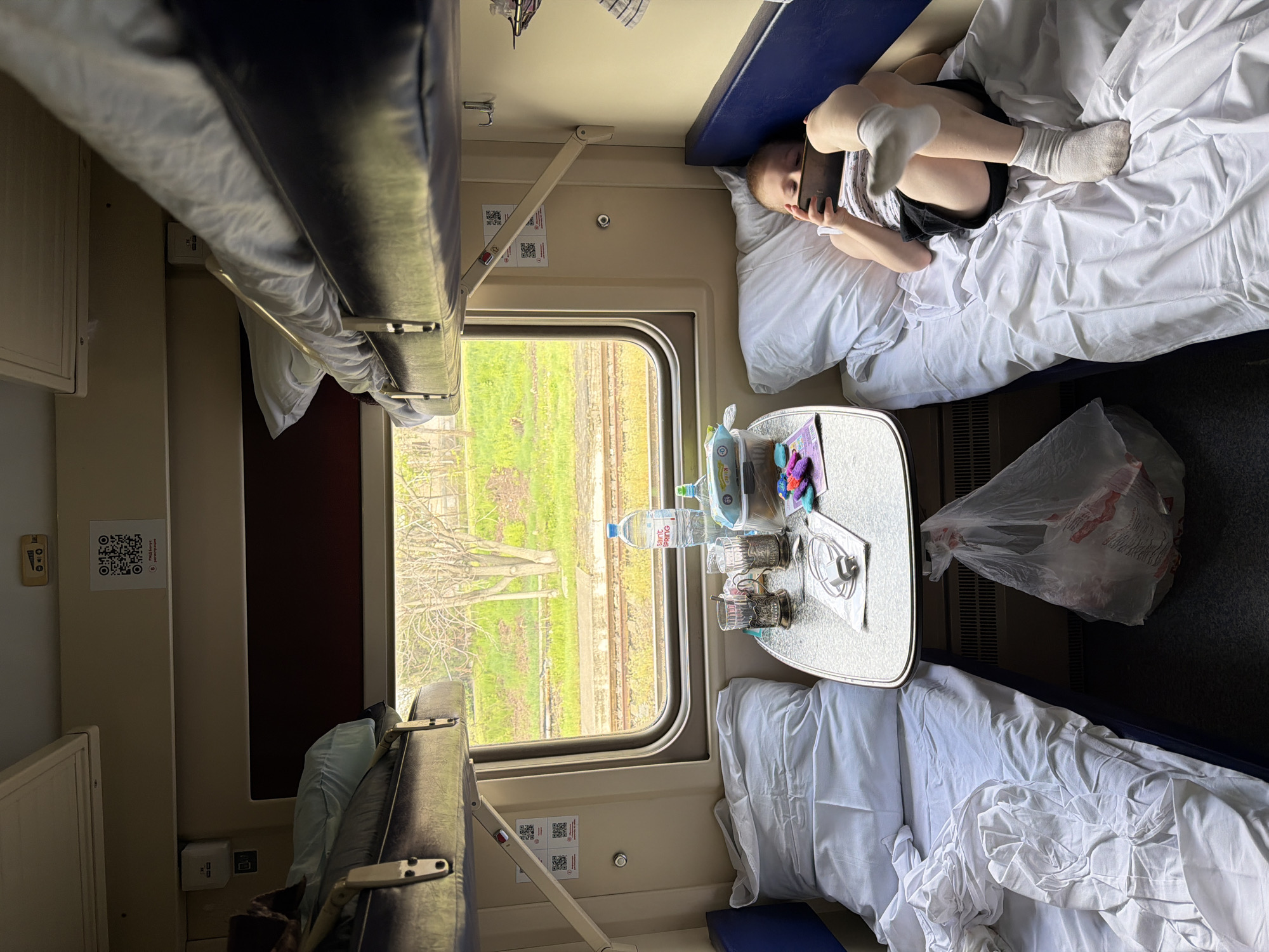 A passenger train interior with two bunk beds, one occupied by a person looking at a phone while reclining. The beds are made with white sheets. A small table between the beds holds drinks, bottles, and other small items. A window shows the blurred view of passing landscape, including trees and railway tracks. The scene shows a typical train journey compartment. While Krasnodar, Russia may be where the photographer is from, this image does not definitively reveal that as a location.