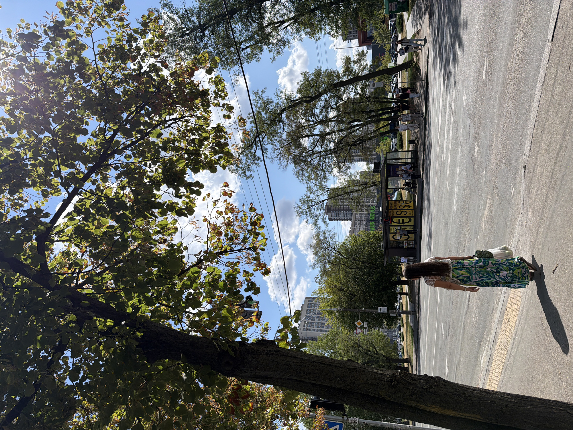 The image depicts a street scene in Kyiv, Ukraine, under a bright, sunny sky with scattered clouds. In the foreground, a woman with long, dark hair, wearing a white lace top and a long, flowing skirt with a green and white floral pattern, walks away from the camera on a paved sidewalk. She carries a green patterned bag and a white plastic bag. A large tree with green leaves partially shades the left side of the image, its trunk prominent and textured.
Across the street, there is a bus stop shelter with yellow advertising panels featuring currency symbols ($, €, £). Several people are visible sitting and standing at the bus stop. Beyond the bus stop, more trees and apartment buildings are visible, some with a modern architectural style. A crosswalk is marked on the asphalt road. Several cars are parked along the roadside in the background. The overall atmosphere is that of a warm, urban day.