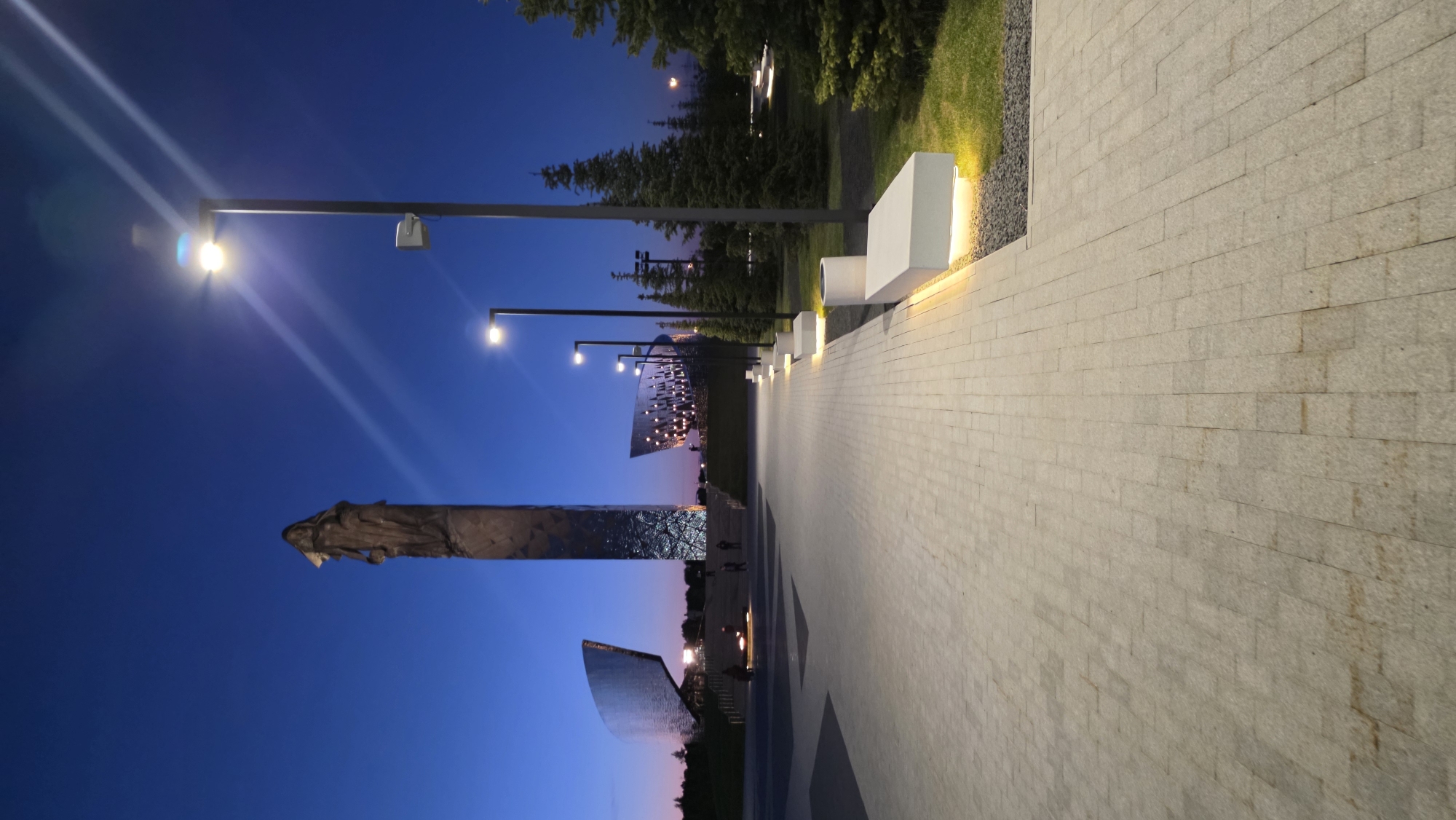 The image shows a paved walkway in Zaitsevo (Verevskoe S/P), Russia, at dusk or night. The walkway is constructed of light-colored rectangular stones, laid horizontally. Parallel to the walkway are regularly spaced benches or light fixtures. To the left of the walkway is a monumental sculpture or structure reflecting the evening sky, consisting of a central high structure with a statue or figure at its top. There are two curved mirror-like structures. Streetlights illuminate the walkway, casting bright light. Trees and vegetation border the walkway on the right, with a section of grass between the walkway and the greenery. The sky transitions from blue to a subtle pink or orange hue near the horizon. There are people walking along the walkway.