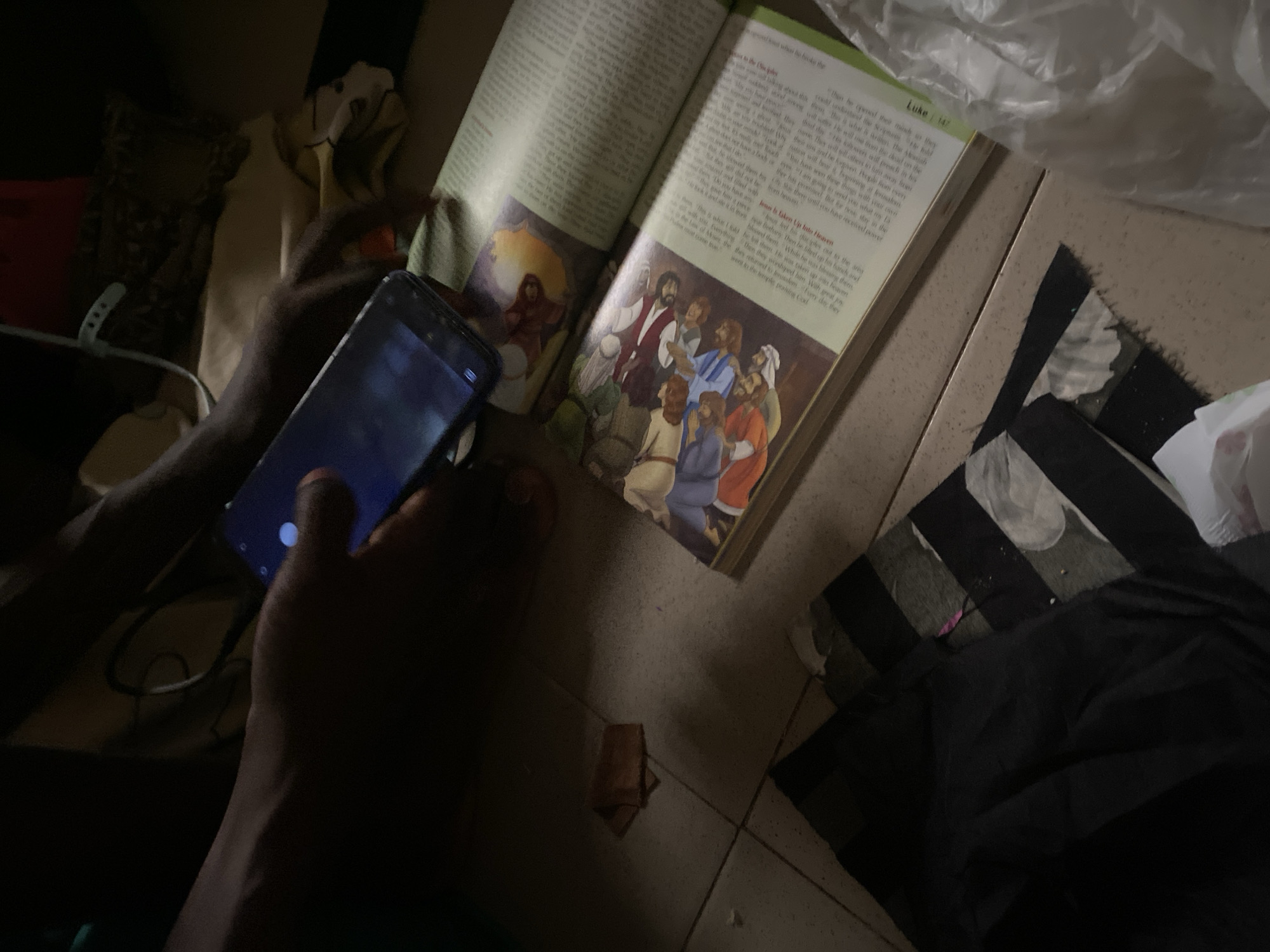 The image depicts a close-up, dimly lit scene, possibly indoors, where a person's hands are interacting with a smartphone that is resting on an open book. The book appears to be an illustrated religious text, with a drawing of a group of people in historical attire on the left page and text on the right page. A charging cable is visible, connected to the smartphone. In the background, there are dark fabrics and what looks like a tiled floor. The scene is set in Jauro Yinu, Nigeria, where the lighting conditions are poor, suggesting it might be nighttime or an area with limited artificial light.