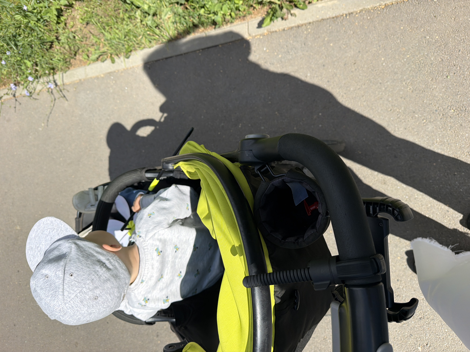 A baby wearing a gray baseball cap and a gray long-sleeved shirt with a pattern of small yellow and blue birds is sitting in a black stroller with a bright green interior. The stroller is on a gray paved walkway, next to a strip of green grass with small purple flowers. The sun is casting a long shadow of the stroller and an unseen person onto the walkway. The scene appears to be a sunny day in Votkinsk, Russia.