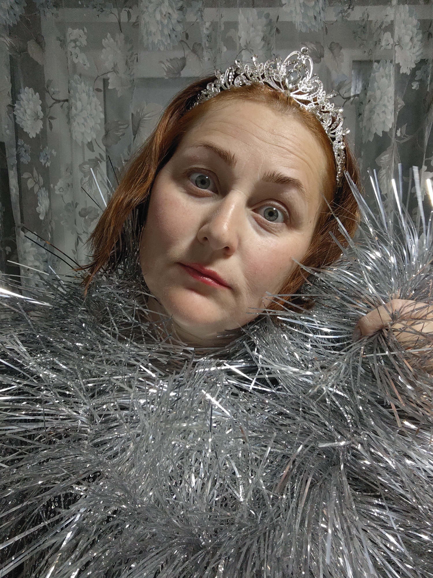 The image shows a woman with red hair and a silver tiara. She is surrounded by silver tinsel. The background appears to be a patterned curtain. The location, Марковский, Russia, cannot be verified from the image.