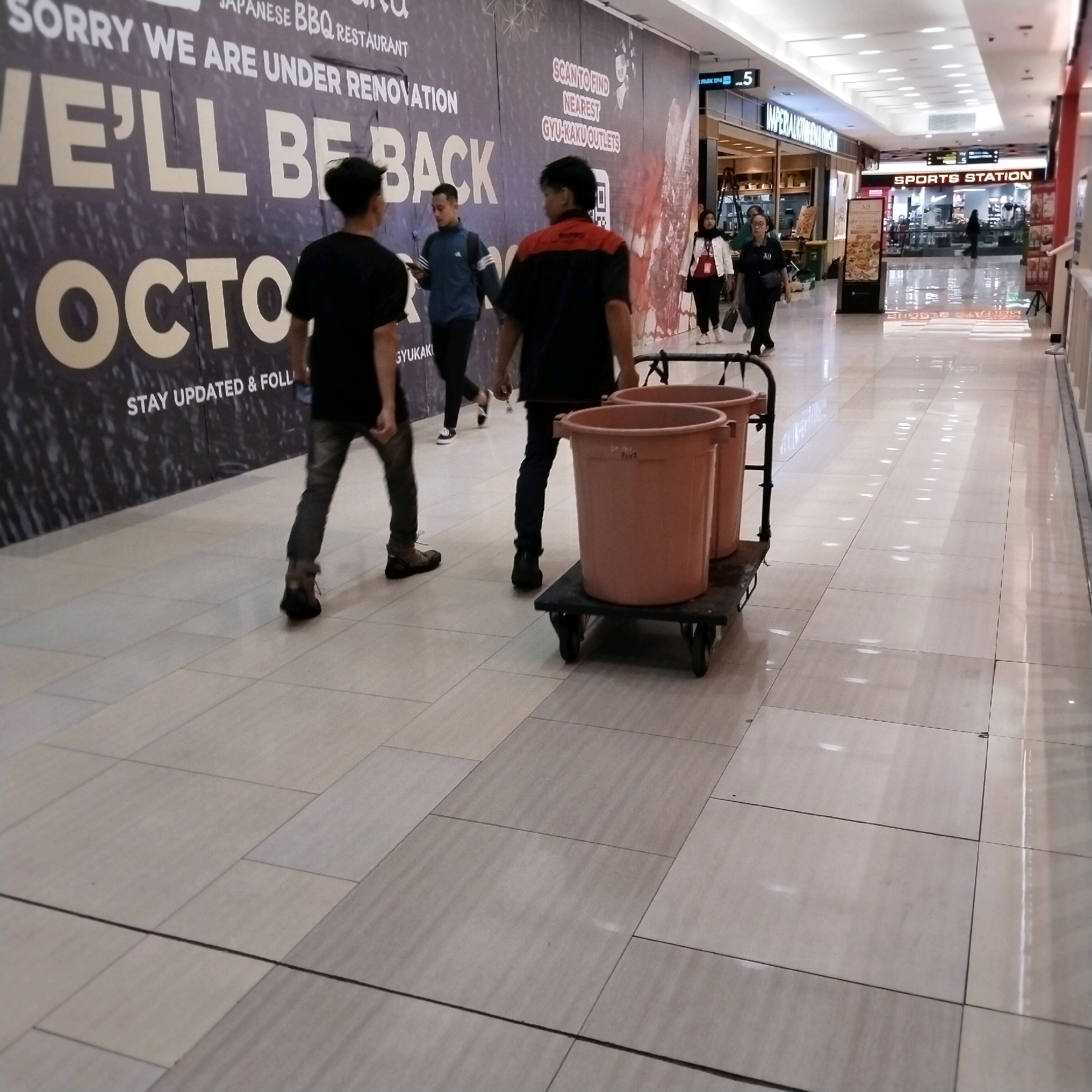In a brightly lit indoor walkway, possibly a shopping mall in Surabaya, Indonesia, two men are walking away from the camera. The man on the left wears a black t-shirt and dark pants, his back to the viewer. The man on the right wears a red and black uniform shirt and dark pants, also with his back to the viewer. They appear to be walking past a large dark banner with white text that reads "SORRY WE ARE UNDER RENOVATION WE'LL BE BACK OCTOBER". In the foreground, two large, light brown plastic buckets are positioned on a dark, wheeled cart. The floor is tiled in a light beige color. Other shoppers and storefronts are visible in the background.