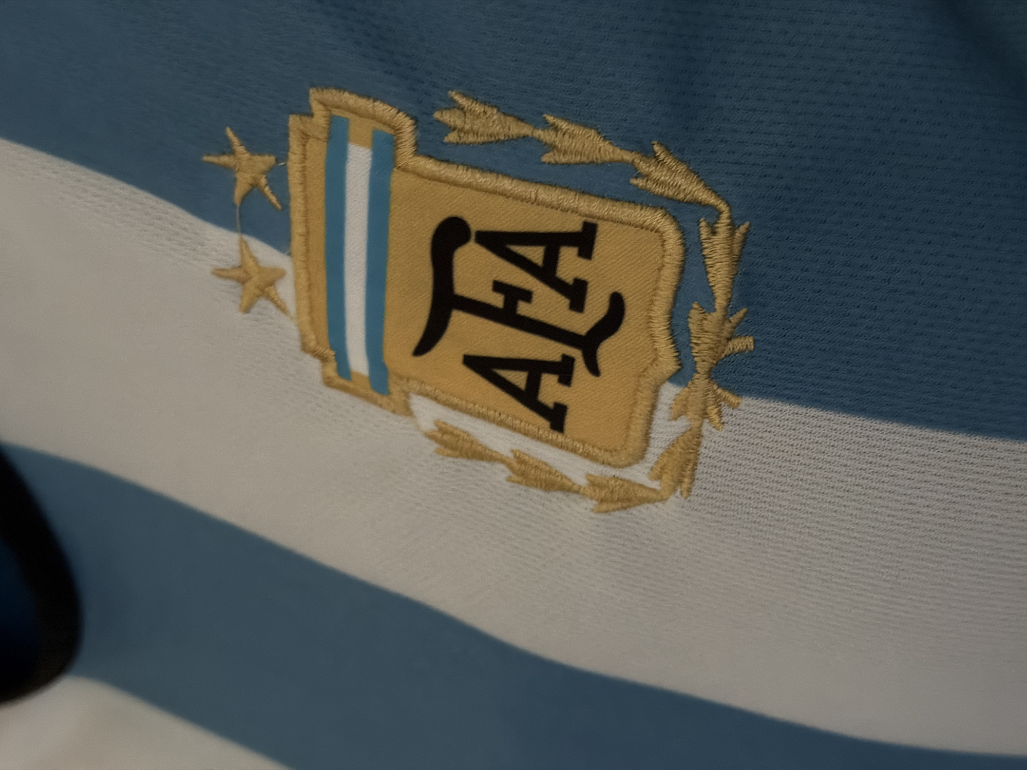 A close-up image showcases an embroidered emblem on a striped fabric, likely a sports jersey or flag, displaying the national colors of Argentina: light blue and white. The central feature is the gold-colored emblem of the Argentine Football Association (AFA). Within a shield shape, the bold black letters "AFA" are visible against a golden background, accompanied by vertical light blue and white stripes on the left side of the shield.
To the left of the shield, two golden embroidered stars are prominent, traditionally symbolizing significant achievements like World Cup victories. The entire emblem is intricately detailed with golden laurel branches encircling parts of the design. The fabric itself exhibits a fine, textured weave, typical of athletic apparel. A notable diagonal shadow cuts across the lower portion of the image, falling over both the white and light blue stripes, suggesting specific lighting conditions. The image emphasizes the detailed embroidery and patriotic design.