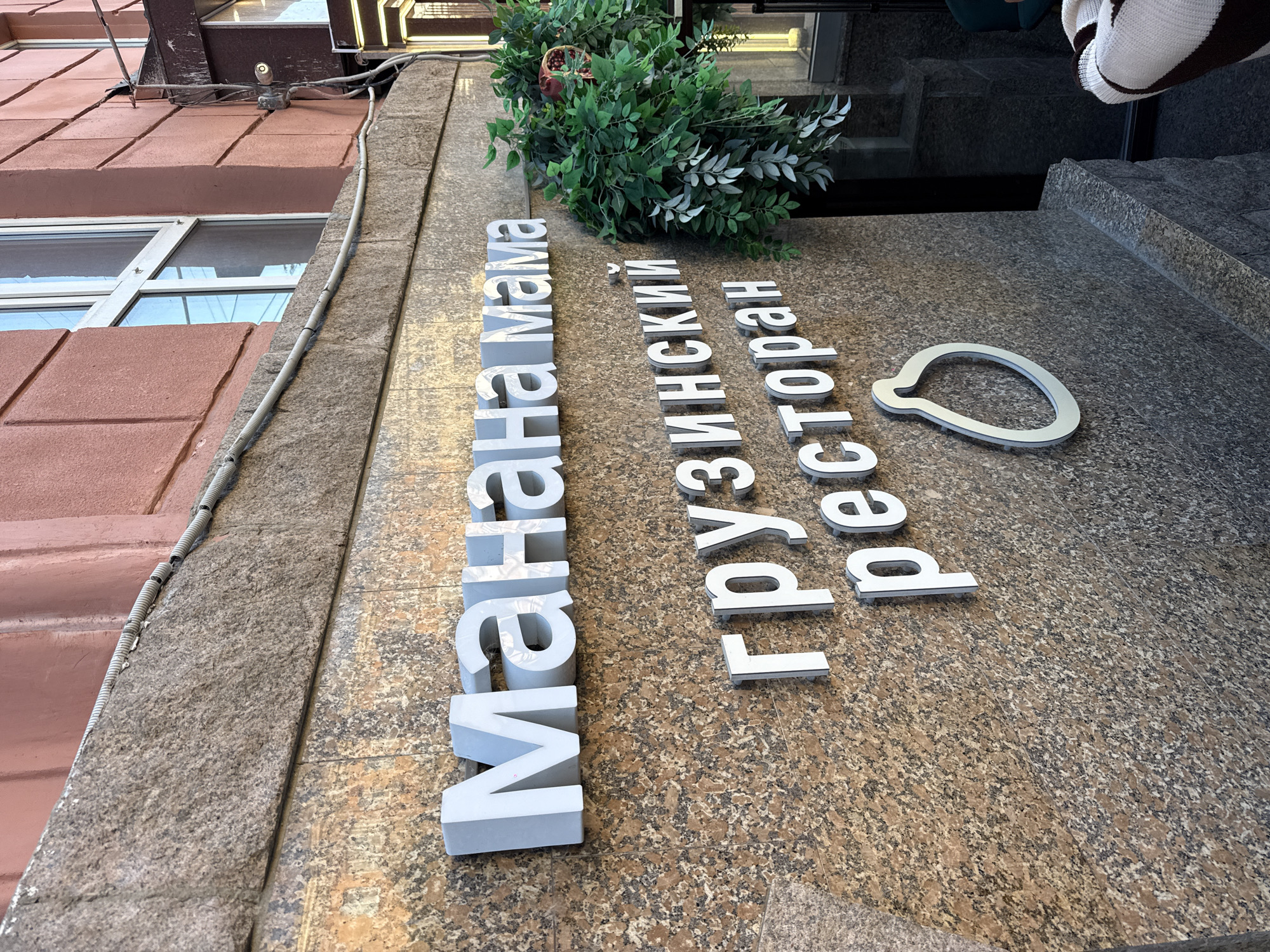 The image shows a close-up of a restaurant's exterior signage. The sign has white, three-dimensional letters spelling "Манана Мама" (Manana Mama) on the top line, and "грузинский ресторан" (Georgian restaurant) on the second line. Below the text, there is a stylized white outline of a pear, which is a common symbol for Georgian cuisine. The signage is mounted on a wall made of speckled granite. To the right of the sign, there is a lush green artificial plant with some leaves appearing to be silver. Part of a doorway and a window are visible to the right, as well as a portion of a person wearing a striped sweater. The building facade above the granite is a reddish-brown color. The overall impression is of a modern and inviting establishment. The restaurant is located in the city of Chelyabinsk, Russia.