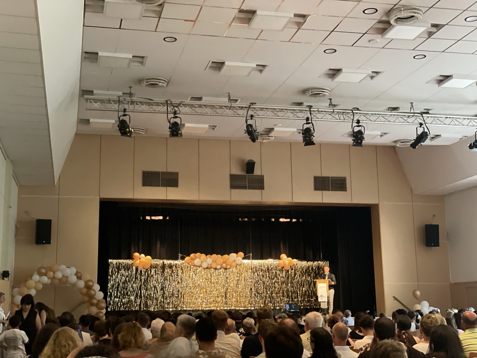 The image depicts an indoor event, possibly a graduation or awards ceremony, taking place in a hall. In the background, a stage is set with a dark curtain, decorated with a shimmering gold fringe backdrop. A cluster of balloons, predominantly white and orange, is arranged across the top of the fringe. To the right of the stage, a man in a dark suit and light trousers stands behind a podium, addressing the audience.
The audience is seated in rows, facing the stage. The hall has a neutral-toned wall paneling. Above, the ceiling features a grid of acoustic tiles, with recessed lighting fixtures and air vents visible. Several stage lights are suspended from a metal truss system. On the left side of the hall, near the entrance or a side aisle, a balloon arch of white and gold balloons is set up. On the far left, a black speaker is mounted on the wall.
The overall atmosphere appears celebratory, with the balloons and the shiny backdrop contributing to a festive ambiance. The image is captured from within the audience, looking towards the stage, providing a perspective of someone attending the event. The event is located at the Stadthalle Nidda, located in Nidda, Germany.