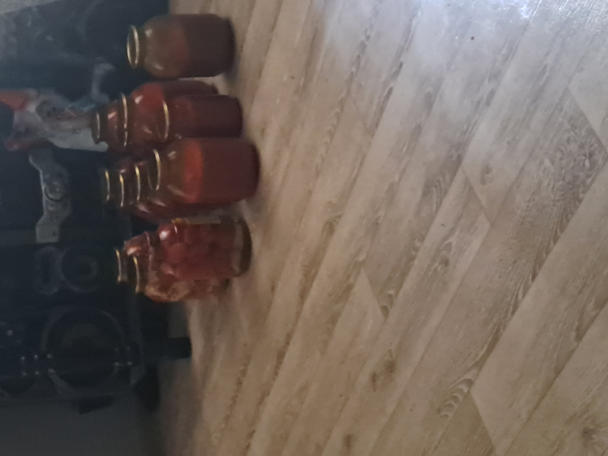The image shows a collection of glass jars containing red preserves, likely tomatoes or berries, placed on a wooden floor. The jars have gold-colored lids. In the background, a dark, large object, possibly furniture or an appliance, is visible. The floor appears to be laminate or wood planking with a light brown hue and a diagonal pattern. The scene is dimly lit. The location is identified as Қарақыстақ ауылдық округі, Kazakhstan.