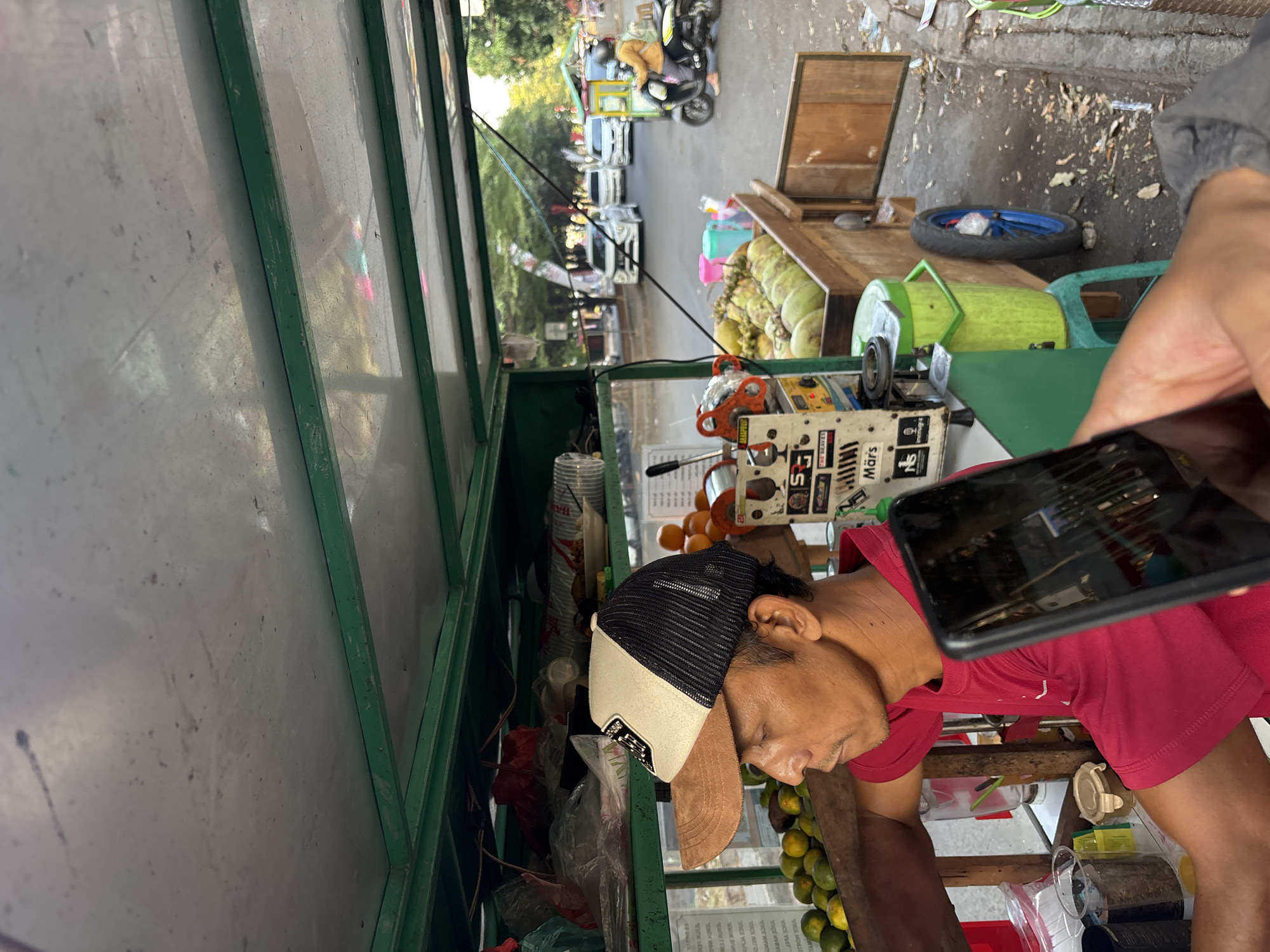 A man wearing a red shirt and a baseball cap is standing at a food cart in Cirebon, Indonesia. The cart is filled with coconuts and oranges. In the background, cars and a motorcycle are visible on a street. The scene appears to be outdoors, possibly in a market or along a busy road. The man is looking to the left, and a smartphone is being held up in the foreground, obscuring part of his face and the cart. The lighting suggests it is daytime, possibly late afternoon.