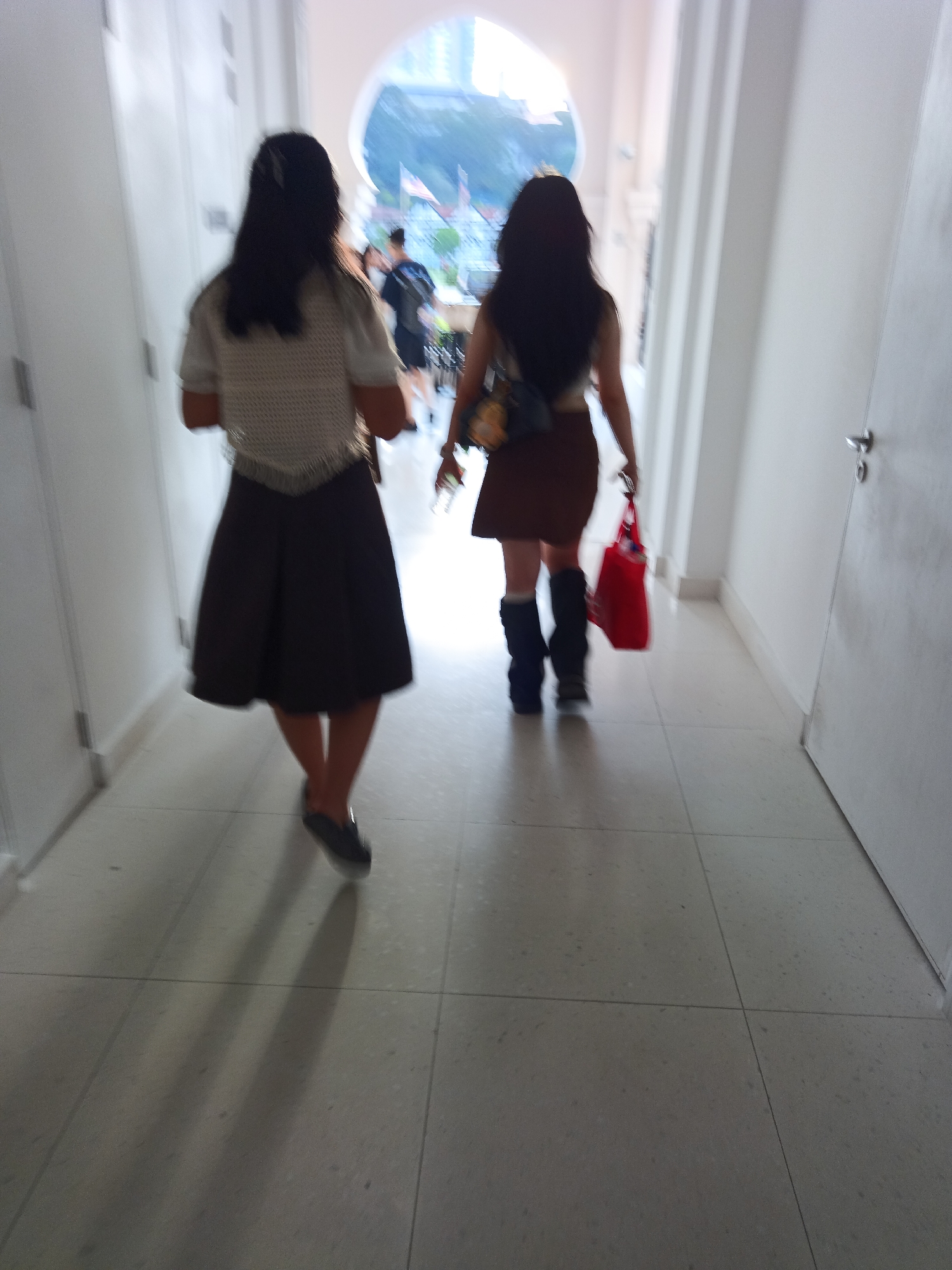 Two individuals, appearing to be young women, walk away from the camera down a light-colored tiled corridor. The corridor has white walls on either side. The individual on the left wears a white, patterned top and a dark skirt. The individual on the right wears a brown skirt and dark boots, and carries a bright red bag. In the distance, an arched opening reveals a glimpse of the outdoors with some structures and flags visible. This scene appears to be inside a building in Kuala Lumpur, Malaysia.