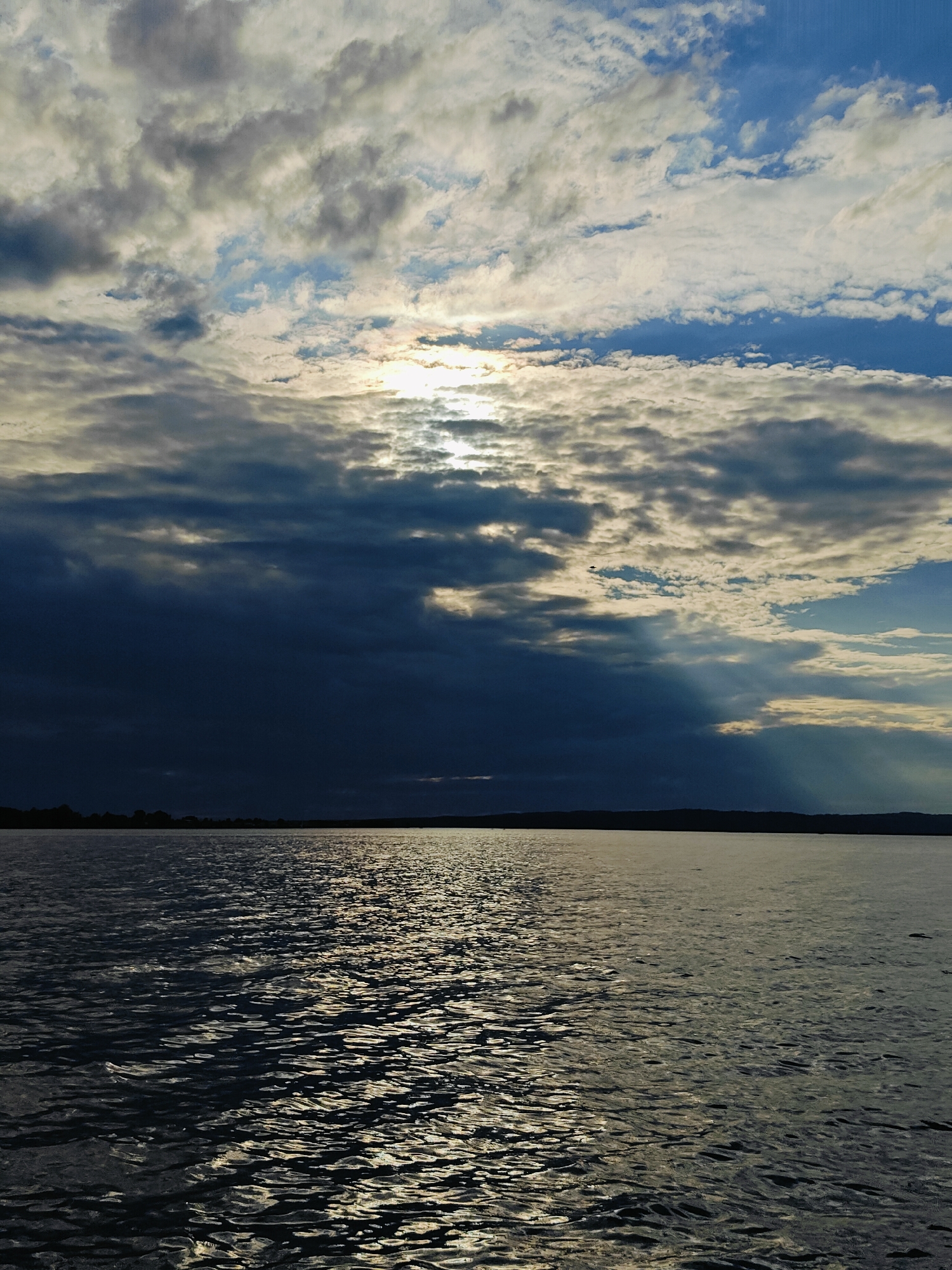 The image depicts a body of water, likely a lake or a large river, with a silhouetted treeline on the horizon. The sky above is dramatic, filled with a mix of dark, heavy clouds and lighter, scattered ones. Rays of sunlight pierce through the clouds, creating shafts of light that illuminate parts of the water's surface and the sky. The water itself is rippled, reflecting the light from the sky and showing a pattern of light and shadow. The scene suggests a moody, possibly transitional weather condition, with the sun breaking through after or before a storm. The overall impression is one of natural beauty and the power of weather. The time of day appears to be either early morning or late afternoon, given the angle of the sun and the quality of the light. There are no visible people or objects in the foreground, and no text is present in the image. The location context of Chaikovsky, Russia, suggests a landscape that could be typical of that region.