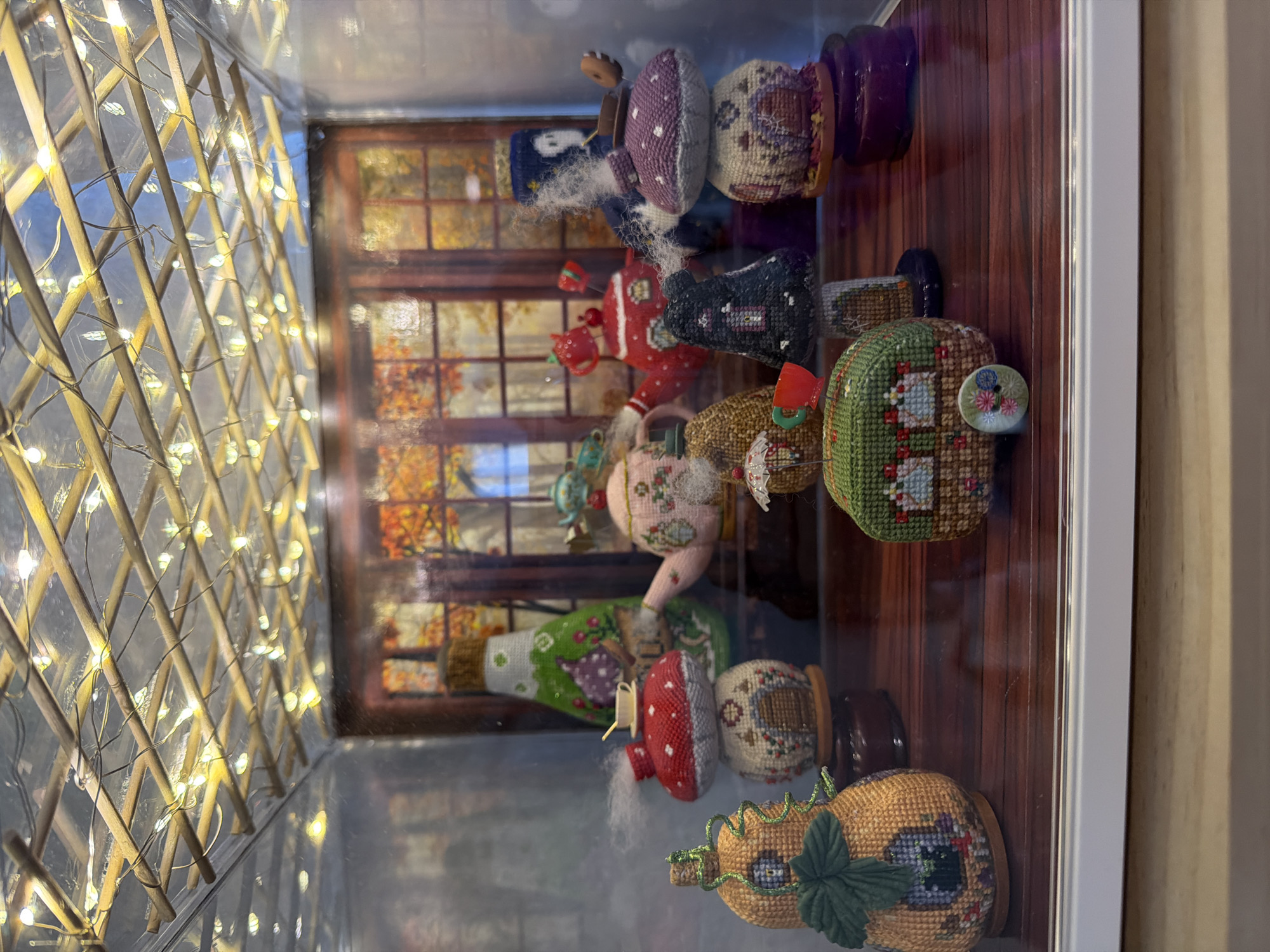 The image shows a diorama with multiple miniature, cross-stitched objects arranged on a wooden surface, enclosed in a glass display case. Above, a lattice of thin wooden sticks is adorned with a string of small, warm-toned LED lights. The backdrop behind the objects appears to be a printed scene of a window with autumn foliage visible through it.
The miniature objects resemble whimsical houses and teapots. On the left, there is an orange, pumpkin-shaped house with green detailing. To its right, a red mushroom-shaped structure sits atop a small house, with a tiny teacup and saucer on the mushroom's cap, and what appears to be wisps of smoke emanating from it. Further to the right, a pink floral teapot is visible, with a small red teapot and cup nearby. In the center foreground, a green, rounded object with a small umbrella and a red cup and saucer sits on a patterned base. Behind this, a black mushroom-shaped house with windows is placed on a dark base. To the right of the black mushroom house, a purple mushroom-shaped house is stacked on top of another house, with a small donut-like object on top. To the far right, a tall, green bottle-shaped object with floral patterns is visible. The overall scene has a fairy-tale or enchanted forest theme.
The image was taken in Aliso Viejo, United States of America.