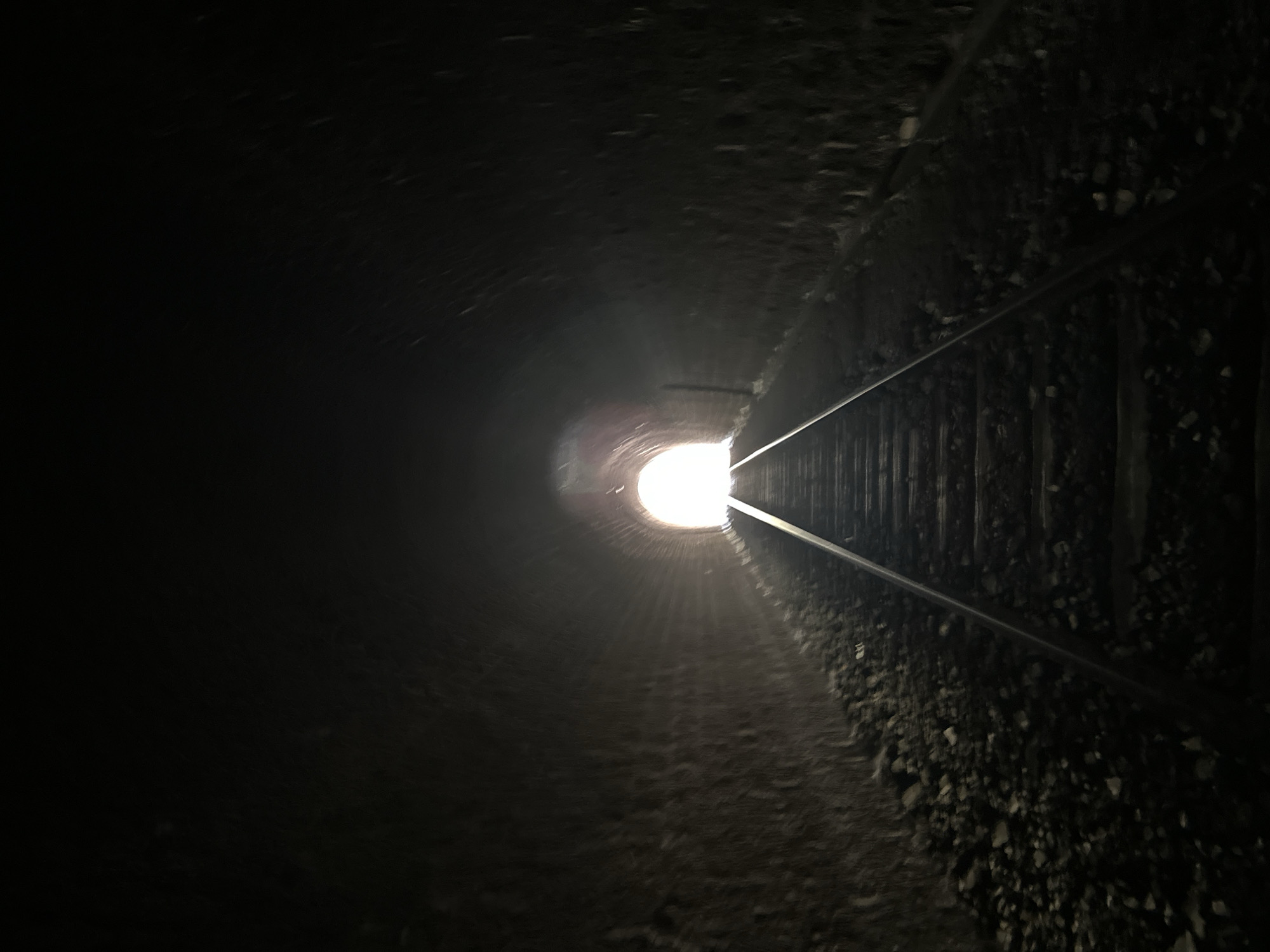 An interior view from inside a dark train tunnel shows a bright light at the end of the tunnel. Railroad tracks lead into the tunnel from the foreground, with ballast and gravel on either side of the tracks. The tunnel walls are rough and textured, appearing to be made of stone or brick. The scene is dimly lit except for the bright illumination emanating from the tunnel's exit. The perspective draws the viewer's eye towards the light, suggesting a journey or an escape from darkness.