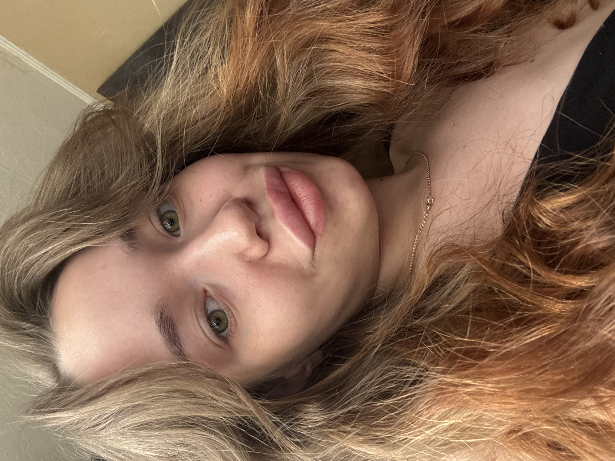 A close-up portrait shows a young woman's face and upper body, with the image rotated 90 degrees clockwise, suggesting she is lying down and looking upwards at the camera. Her head is positioned towards the left side of the frame.
**Subjects:** The primary subject is a young woman.
**Activity or event taking place:** She is posing for a photograph, engaging directly with the camera with a calm and neutral expression.
**Notable details:**
* **Appearance:** She has light skin, distinct green or hazel eyes, and full, naturally pink lips that are slightly parted. Her eyebrows are well-defined. Her long, voluminous hair is a blend of blonde and warm auburn or light brown tones, appearing wavy and framing her face while spreading around her head and shoulders.
* **Accessories:** A delicate gold-colored chain necklace is visible around her neck and clavicle area.
* **Clothing:** A dark, possibly black, garment is visible on her shoulder.
* **Setting:** The background is simple and out of focus, suggesting an indoor, personal setting with light-colored walls or ceiling and a darker, indistinct object partially visible.
* **Lighting:** The lighting is soft and even, illuminating her features without harsh shadows.
* **Emotions:** Her gaze is direct and contemplative.
* **Time of day:** The time of day cannot be determined from the available light.
* **Location cues:** There are no specific visual cues within the image to indicate the location as Chaikovsky, Russia.
**Visible text:** There is no visible text in the image.