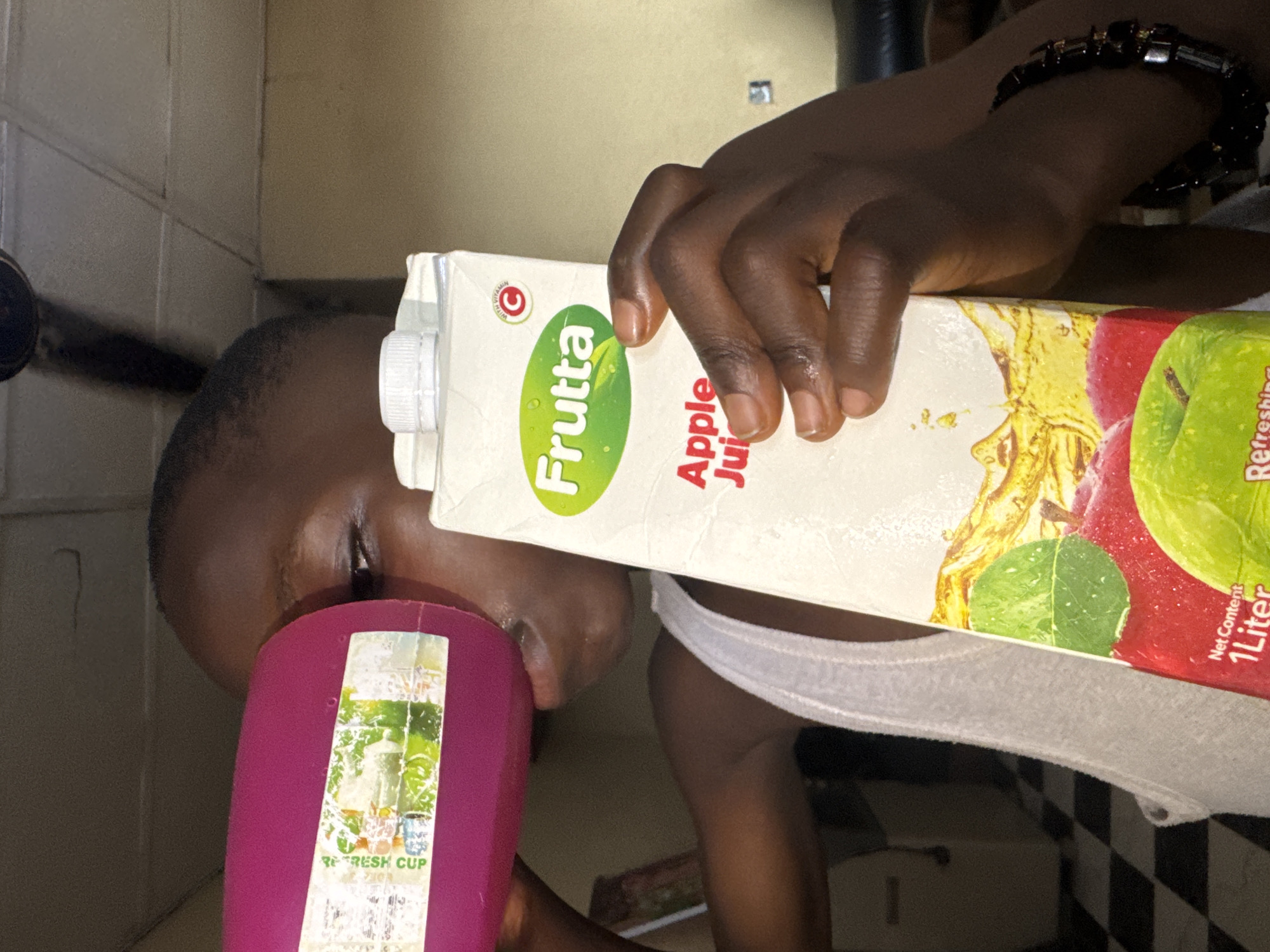 A person is drinking from a pink cup, pouring apple juice from a Frutta brand carton into it. The carton indicates it contains one liter. The individual has dark skin, is wearing a white tank top, and has a dark beaded bracelet on their wrist. The scene is indoors, possibly a kitchen, with tiled flooring and beige-colored walls. The specific location within Onitsha, Nigeria, cannot be determined from the image.