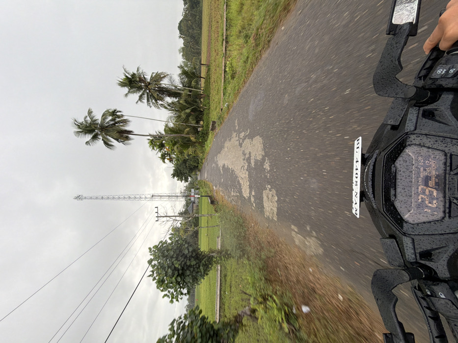 The image depicts a first-person perspective from a moving motorcycle on a wet, paved road in Keudekarieng, Indonesia, under an overcast sky. The black motorcycle's dashboard, handlebars, and a portion of the digital display are visible in the foreground, all covered in raindrops. The display shows "123 Km/h," a time of "6:28," and "245 12" (likely odometer/trip meter). A white license plate, "BG 4408 NAM," is mounted above the dashboard. A hand is visible gripping the right handlebar.
The asphalt road, exhibiting areas of wear and lighter patches, stretches straight into the distance, showing motion blur indicative of travel. To the left of the road, green fields, likely rice paddies, are visible beyond a strip of grass and small trees. Overhead power lines traverse the sky, supported by a wooden utility pole. A prominent, tall, lattice-structured cellular tower stands further down the road on the left, with a building partially visible behind it. To the right of the road, dense tropical vegetation, including several tall palm trees and other broadleaf trees, borders the path, with more green fields visible beyond. The sky is uniformly gray and overcast.