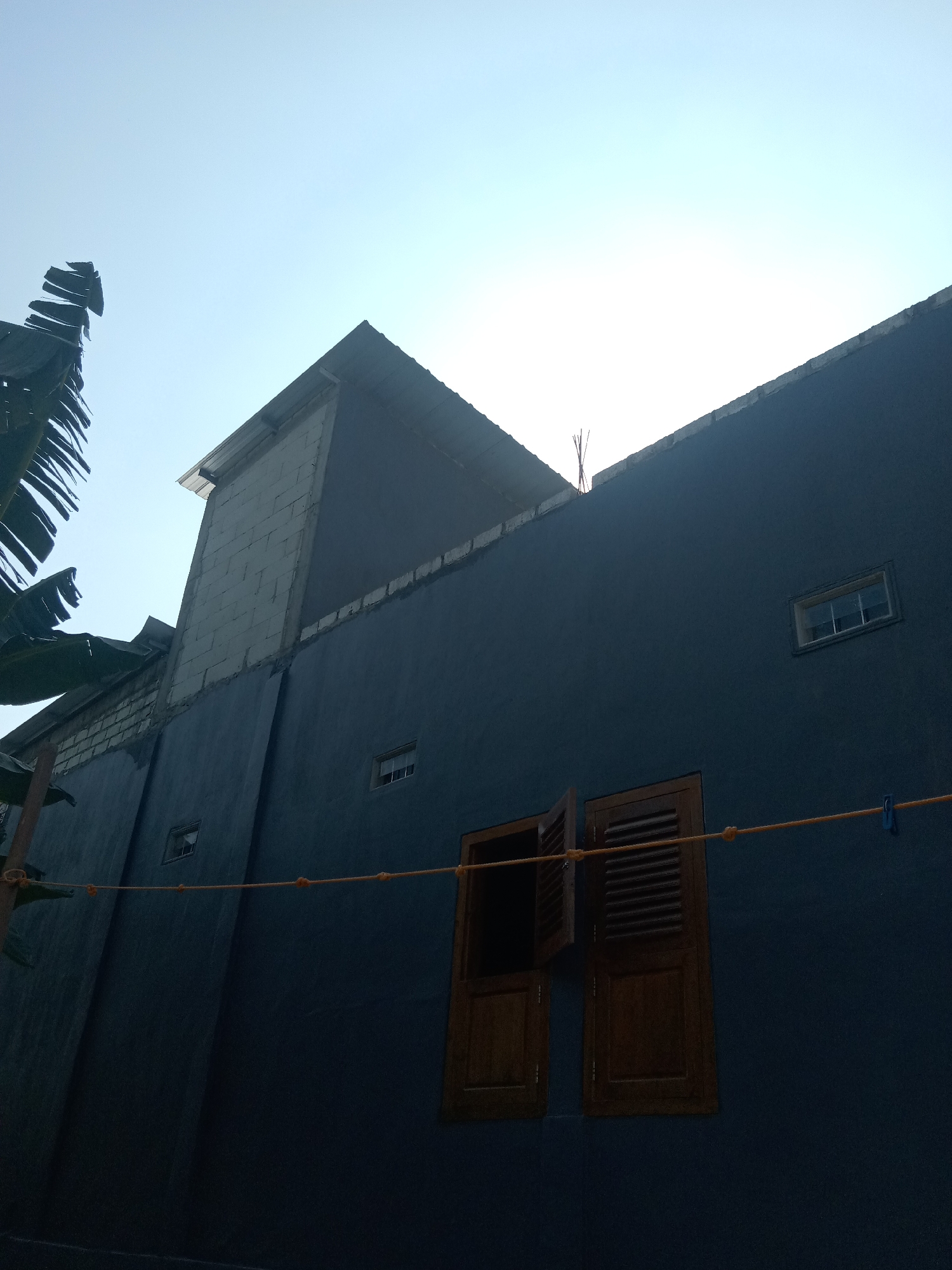 This is a low-angle outdoor photograph of a dark blue building under a clear sky. The building appears to be under construction or renovation, with exposed cinder blocks visible along the top of the dark blue wall, and some exposed rebar protruding from the roofline. There are several windows on the building's facade. Two small, square windows with white frames are visible on the upper portion of the wall, and a larger, wooden-shuttered window is open on the lower portion. A thin orange cord, possibly a clothesline, is strung horizontally across the front of the building, secured by a blue clothespin on the right. Banana leaves from a nearby plant are visible on the left side of the frame. The image is taken in Tegalkodo, Indonesia.
