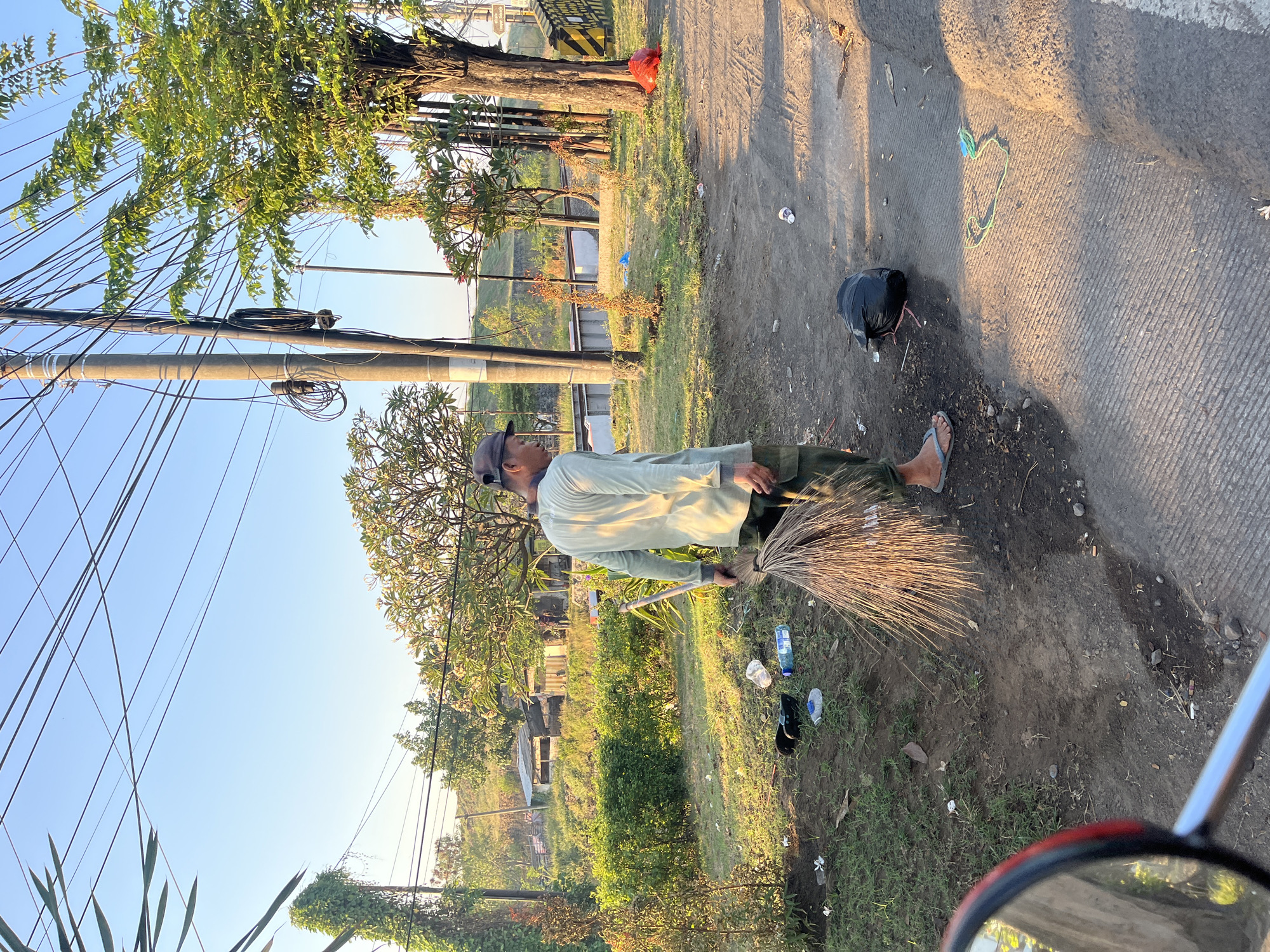 In Kalitengah, Indonesia, a man is sweeping a dirt path with a broom made of palm fronds. He is wearing a light-colored long-sleeved shirt, dark pants, and a cap. A black helmet lies on the ground near him. The area is littered with trash, including plastic bottles, bags, and wrappers. In the background, there are trees, a building with a corrugated metal roof, and utility poles with many wires. The sun is low in the sky, casting long shadows. The scene appears to be on the side of a road, with part of a red side-view mirror visible in the foreground on the left, suggesting the photo was taken from a vehicle.