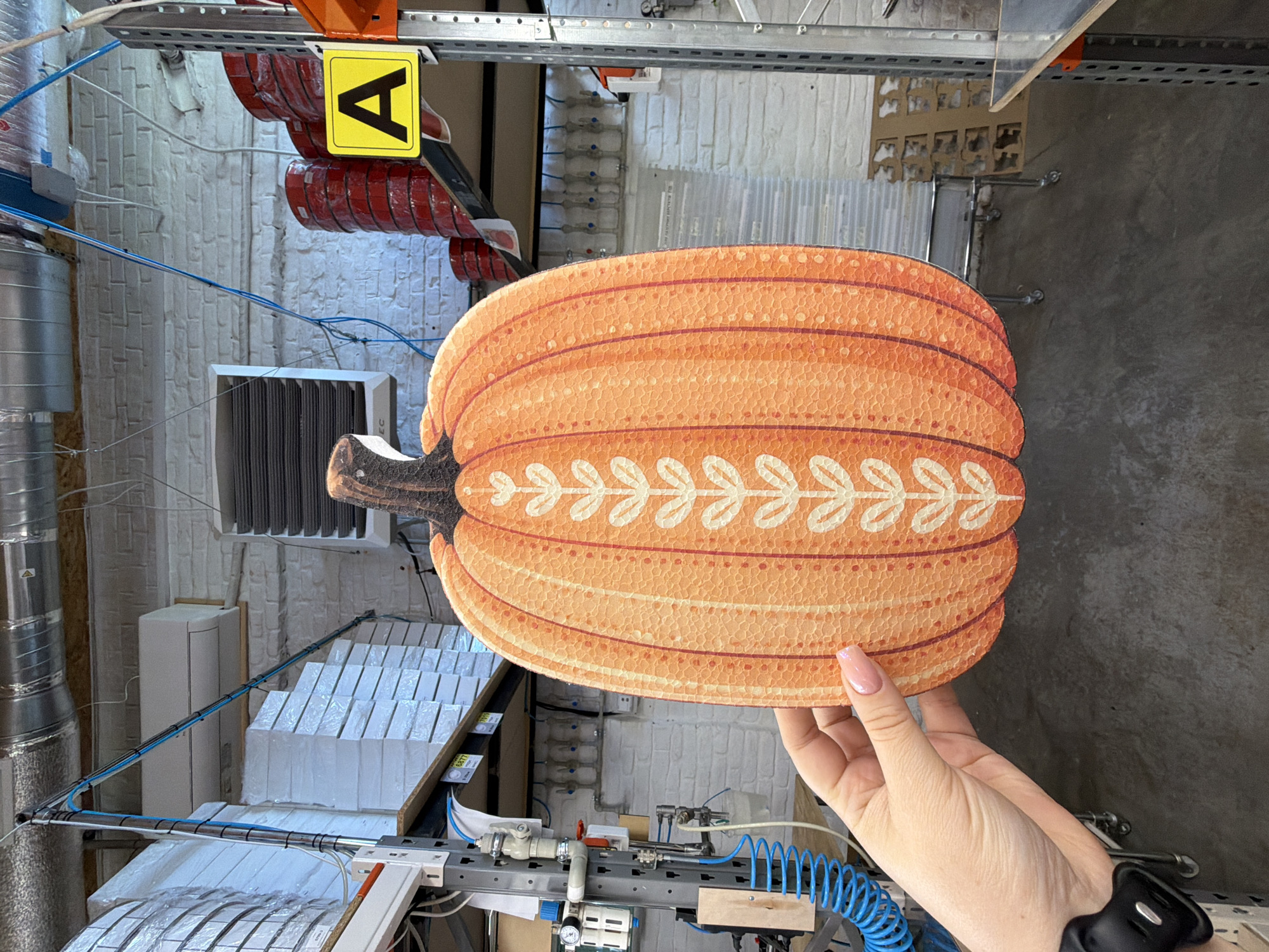 A person's hand with a pink manicure holds a large, orange, pumpkin-shaped cutout with white decorative patterns. The cutout has a textured surface and appears to be made of cardboard. The background suggests an industrial or storage setting, with metal shelving units, stacks of white boxes wrapped in plastic, and air ducts visible on the ceiling. There are also red-colored rolls of material stacked on a shelf, and a yellow sign with a black "A" on it. The floor is a dark, concrete-like material. The lighting is neutral and appears to be overhead.